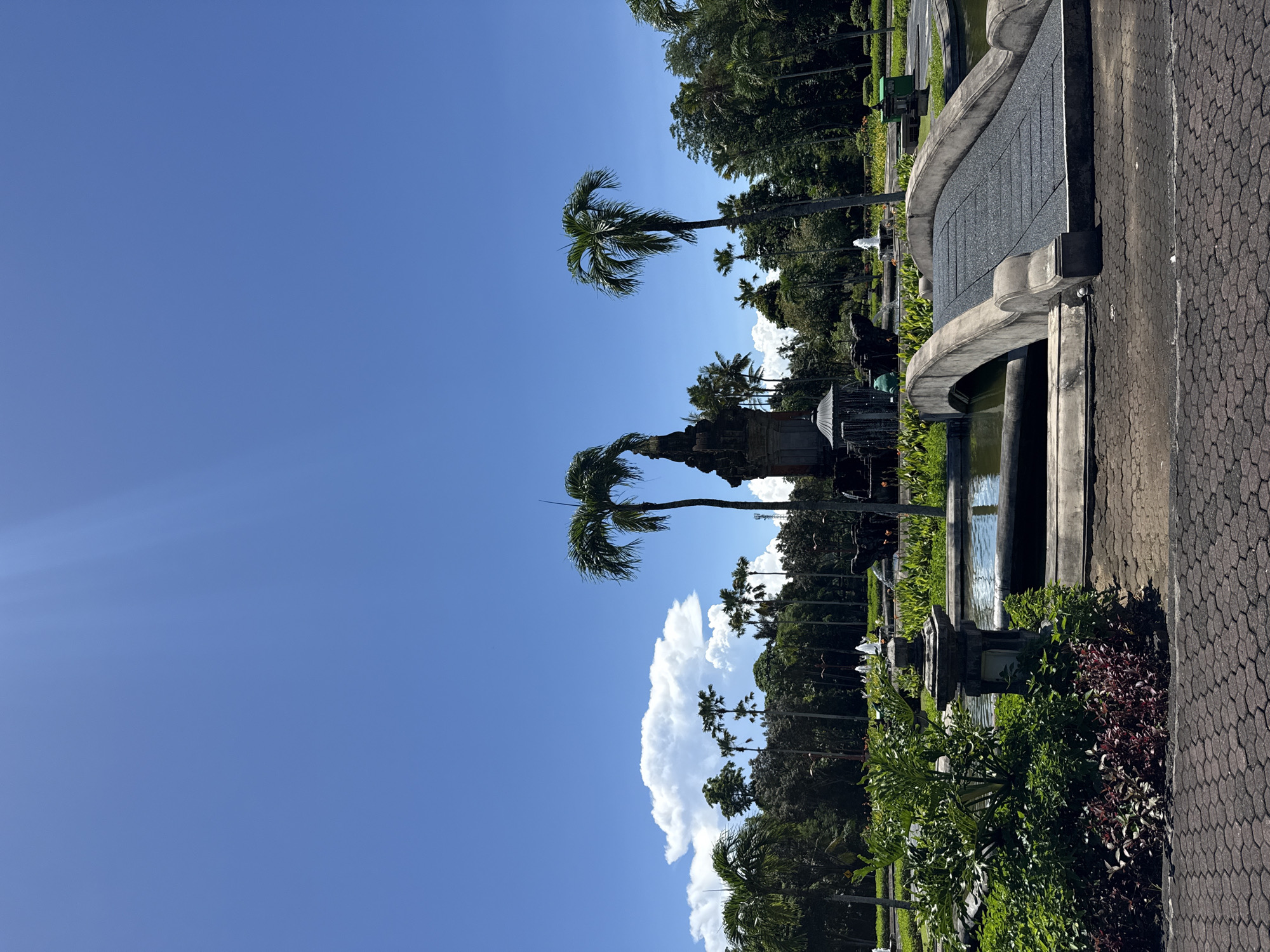 The image shows a park scene in Nusa Dua, Indonesia. The sky is blue with some white clouds. Several palm trees are in the park, and a stone statue is present. There's a decorative concrete water feature with a small pond. A paved walkway runs along the edge of the park. The landscape includes various green plants and shrubs.