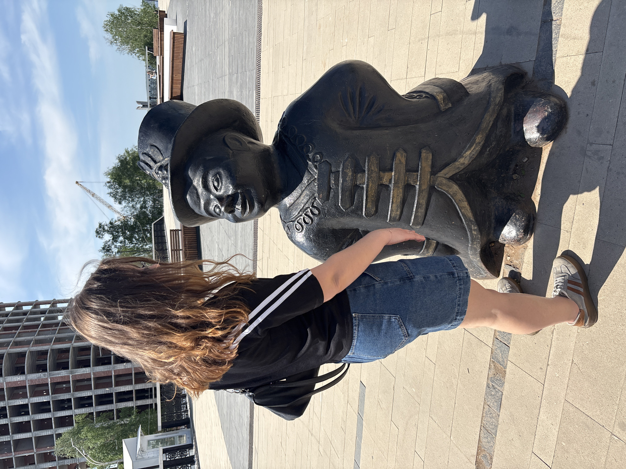 A woman stands on a light-colored brick plaza next to a black metal statue of a man wearing a hat and a coat. The woman has long brown hair and is wearing a black t-shirt with white stripes on the sleeves, a blue denim skirt, and grey shoes with brown stripes. She is facing the statue and has her right hand on its chest. In the background, there is a building under construction and some trees. The image was taken in the city of Izhevsk, Russia.