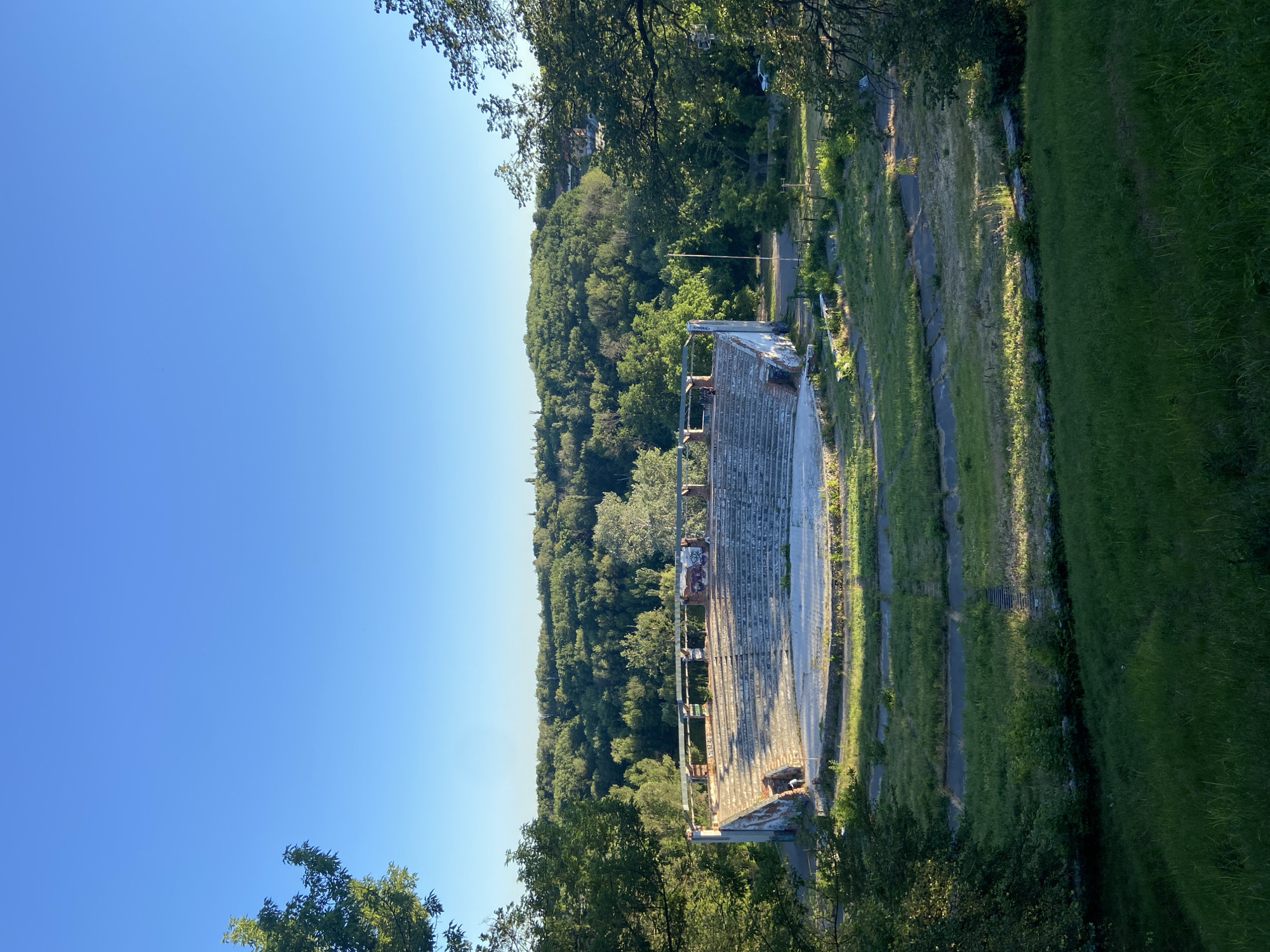 The image depicts an outdoor scene with a clear blue sky. The focal point is a dilapidated grandstand, with rows of weathered seating and exposed metal structures. The grandstand is situated in a grassy, overgrown area, suggesting it is disused or abandoned. Beyond the grandstand, a dense forest covers a hillside that recedes into the distance, dotted with some buildings on the upper slopes. The lighting indicates it is daytime, with the sun casting long shadows across the grass, suggesting it is either early morning or late afternoon. The overall atmosphere is one of decay and nature reclaiming a man-made structure. The image is oriented vertically. While the image is set in Nyzhni Mlyny, Ukraine, there are no specific visual cues within the frame that confirm this location without prior knowledge. There is no visible text in the image.