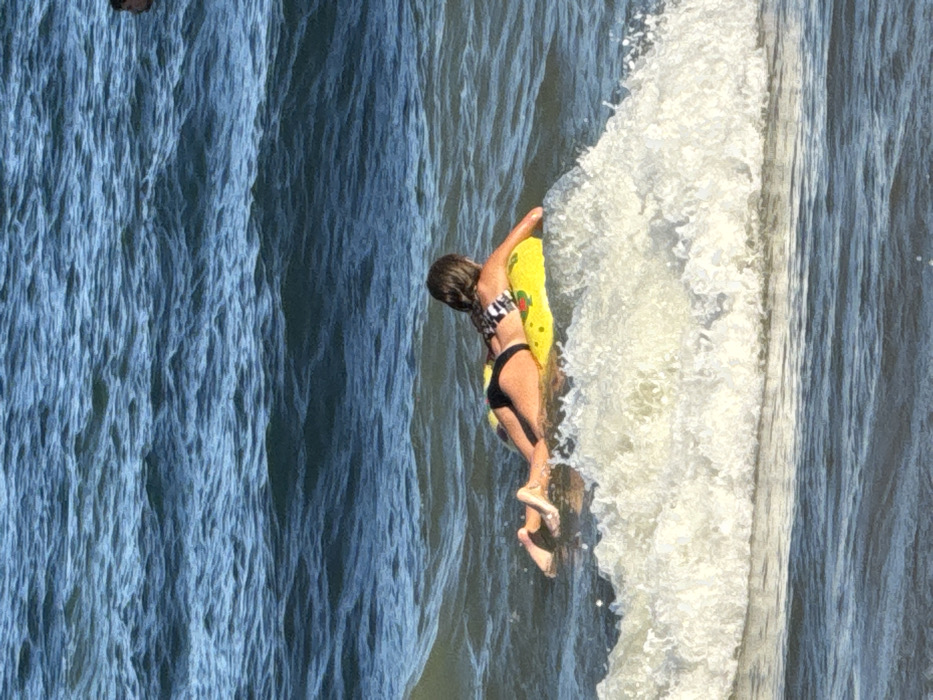 An individual is depicted swimming in the ocean off the coast of Nha Trang, Vietnam, during daylight hours. The subject, appearing to be female with brown hair, is positioned prone on a circular yellow inflatable float featuring dark patterns. She is wearing a black bikini bottom and a black and white patterned bikini top. Her legs are extended behind her. A breaking white wave is visible in the foreground, directly in front of the float. The ocean surface exhibits undulating waves of blue-green water. The head of another individual is partially visible in the upper right background, further out in the water.