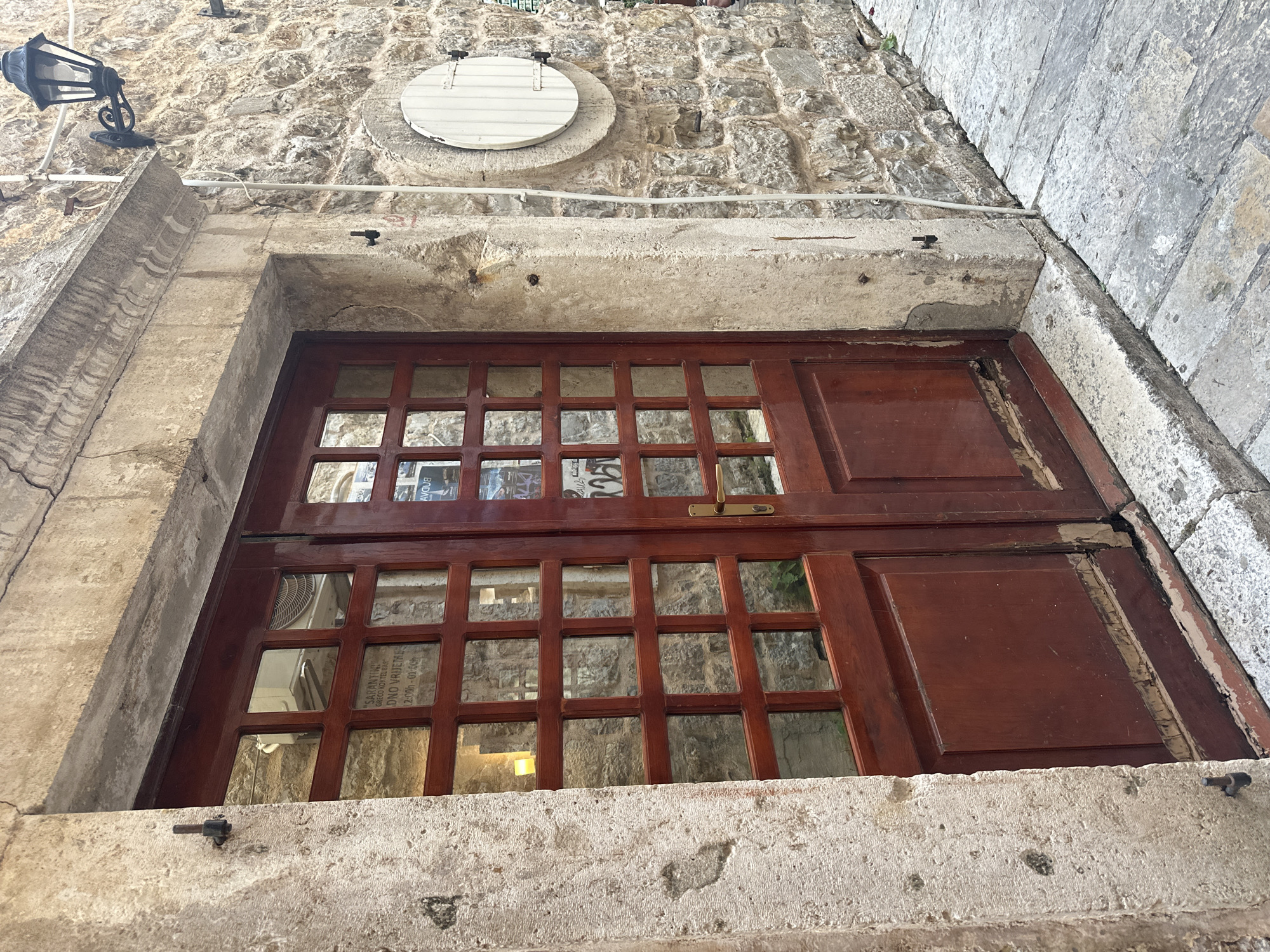 The image depicts a two-part wooden door with glass panes set into a stone wall, likely in an old building. The door is a reddish-brown color and shows some wear. Above the door, the stone wall is textured and aged. There is a round white sign above the door and to the left a vintage-style black metal light fixture is attached to the wall. Through the glass panes of the door, a glimpse of an interior space can be seen, with posters and what appears to be a sign with some text visible. In particular, the text "SARANTI GRECO KOKTEL BAR" and "DNO VRIJEME 12:00-01:00h" is discernable. The setting is likely in the city of Budva, Montenegro.