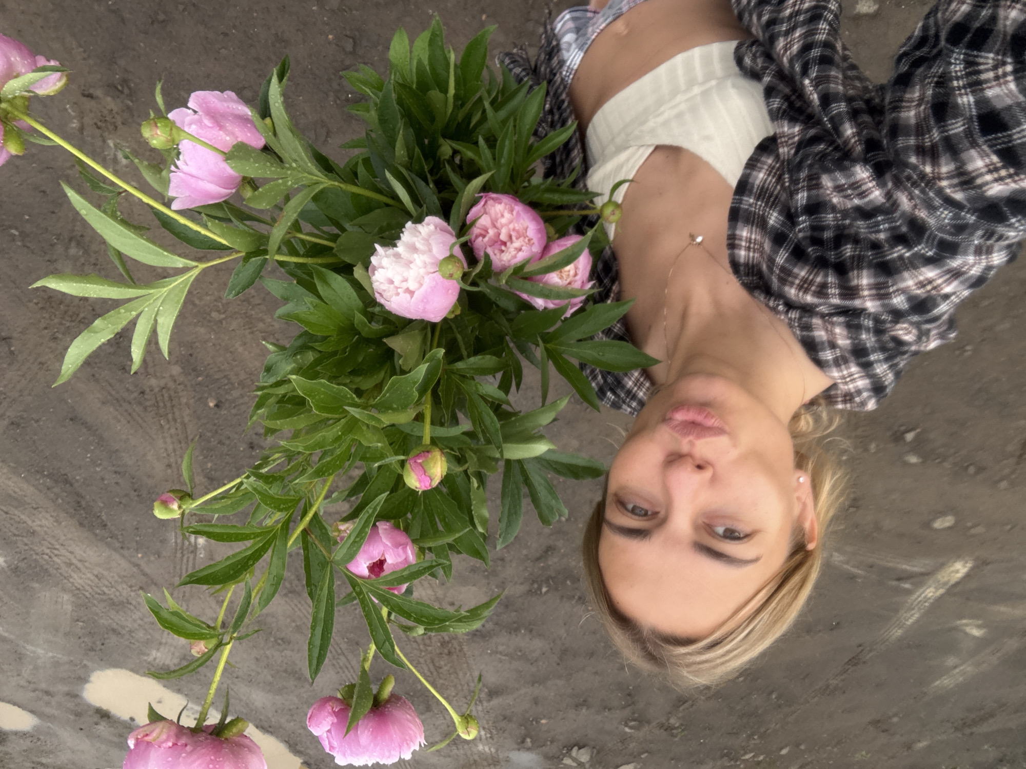 A woman with blonde hair and light skin is taking a selfie outdoors. She is wearing a white ribbed crop top and an open, unbuttoned plaid shirt over it. She is holding a large bouquet of pink peonies, some of which are in full bloom and others are still buds. Water droplets are visible on the flowers and leaves, suggesting it may have recently rained. The woman has her lips pursed as if blowing a kiss or making a kissy face. Her eyes are looking towards the camera. She is wearing a delicate necklace with a small charm. The background is a muddy, uneven ground with tire tracks and some puddles, indicative of an unpaved area, possibly a field or a construction site, that could be found in an industrial or rural part of Ust-Ilimsk, Russia. The overall lighting suggests it is daytime.