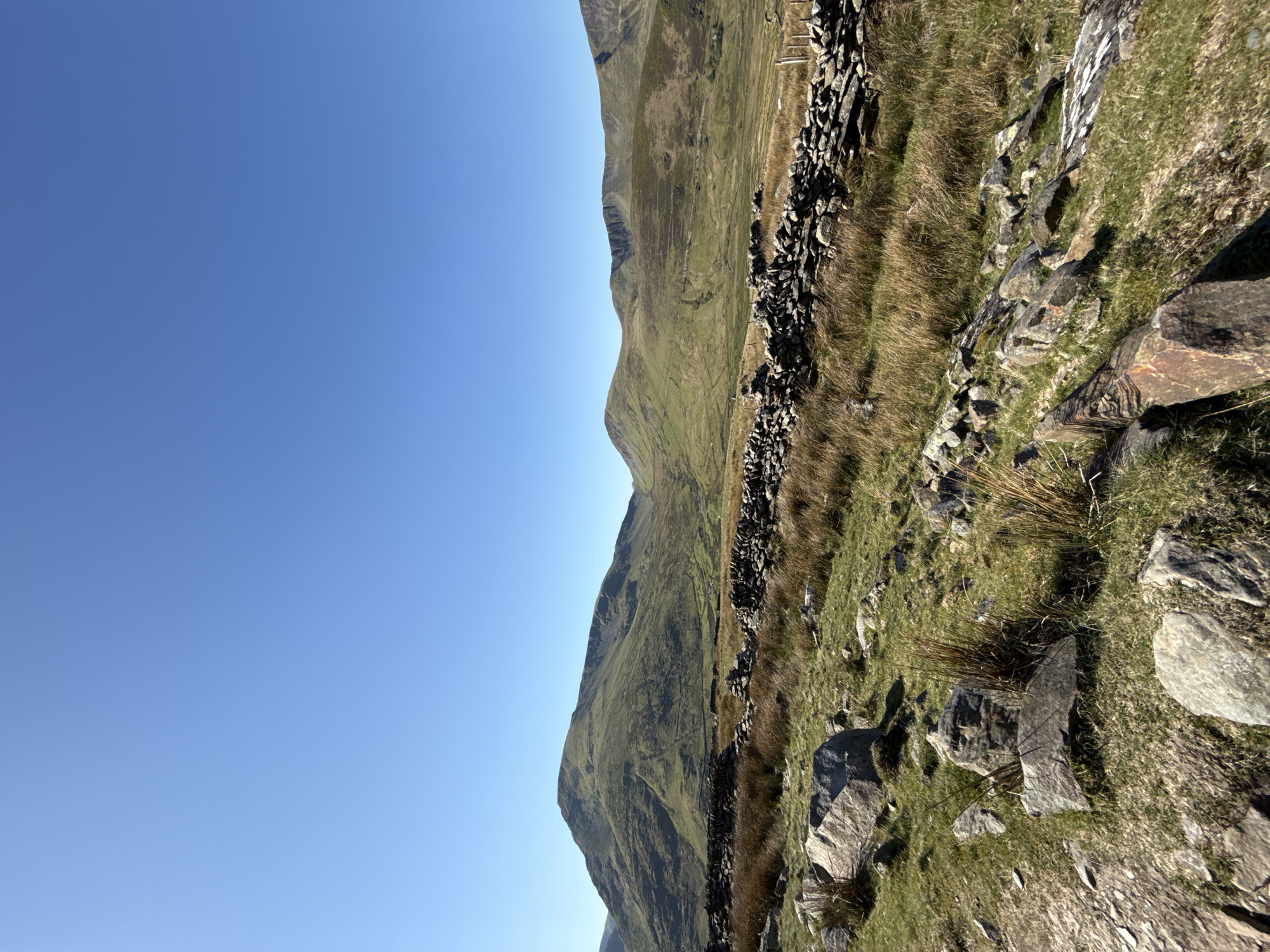 The image depicts a landscape scene in Llanberis, United Kingdom. The composition features a clear blue sky occupying the left portion of the frame. A series of rolling, grassy mountains extends diagonally across the image. A stone wall constructed of dark rocks runs along the slope. Grass, weeds, and additional rocks and boulders are scattered throughout the foreground and middle ground of the landscape. There are no people visible.