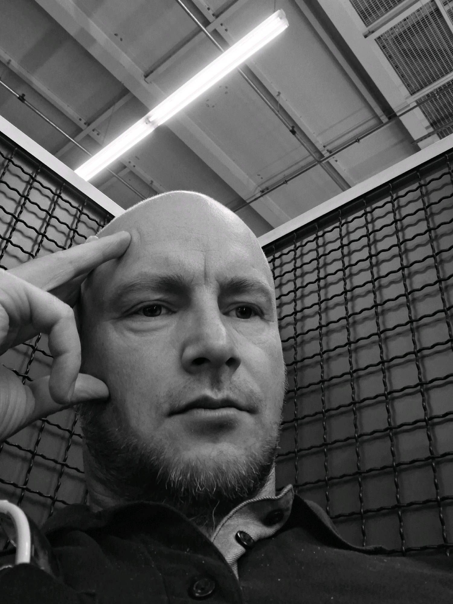 A black and white, close-up, low-angle, head-and-shoulders selfie of a bald, middle-aged white man. The man is looking towards the viewer with his right index finger touching his forehead. He has a light beard and is wearing a dark collared shirt. The background consists of a black wire mesh fence and a ceiling with fluorescent lighting and ventilation systems. The scene appears to be indoors, possibly in a warehouse or industrial setting. The man's expression is pensive.