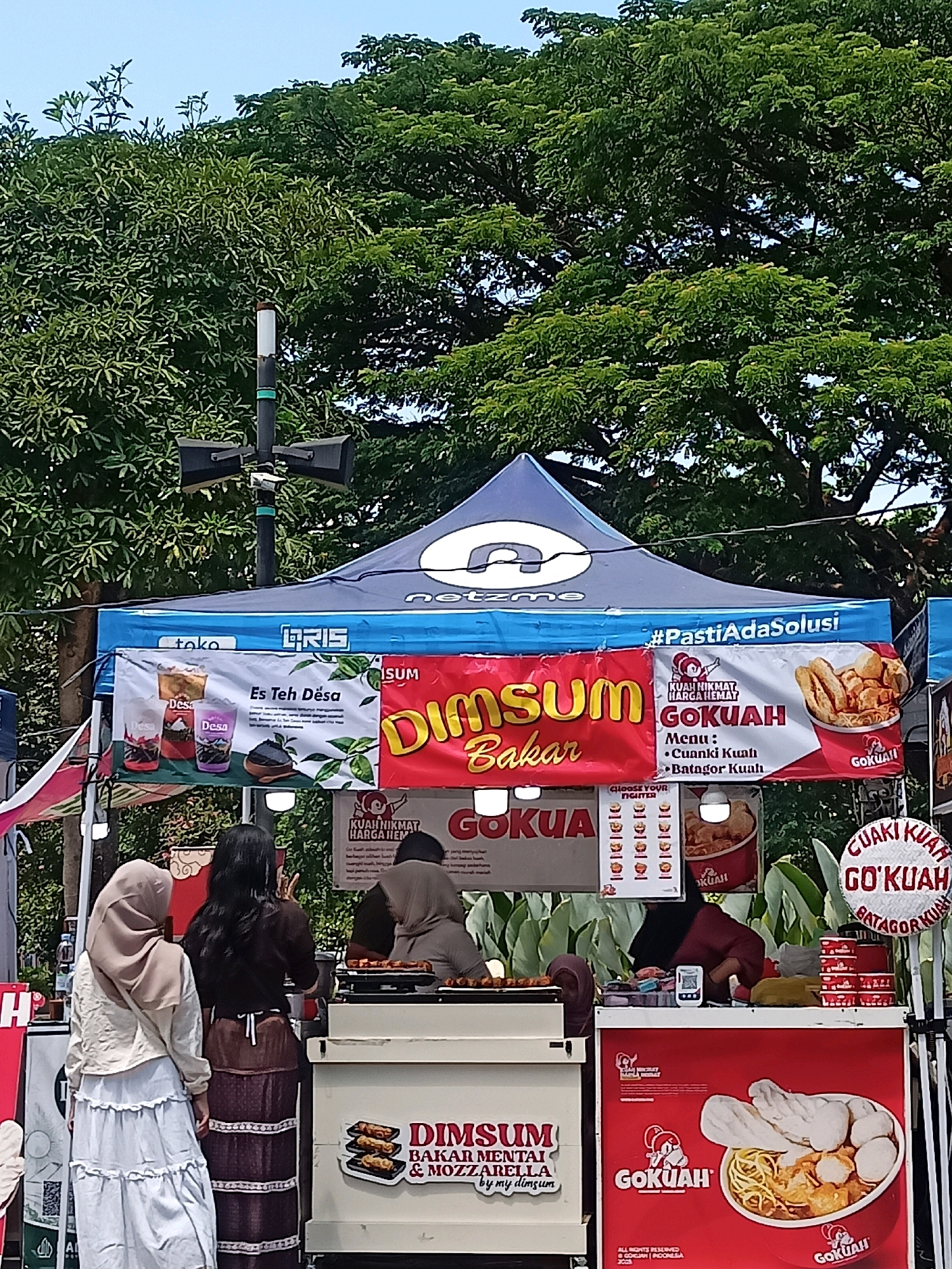 The image shows an outdoor food stall in the city of Surakarta, Indonesia. The stall is set up under a blue and white tent. On the tent's canopy, the text "net" is visible. Below the canopy, there are several banners advertising food items. One banner features the text "Es Teh Desa" with images of drinks. Another banner displays "Dimsum Bakar" and "GOKUAH" with an image of fried food. A third banner promotes "KUAH NIKMAT HARGA HEMAT GOKUAH" and lists menu items: "Cuaniki Kuah" and "Batagor Kuah." A white refrigerator unit displays "DIMSUM BAKAR MENTAL & MOZZARELLA by my dimsum". Another banner on the right shows "GUAKI KUAH GO'KUAH BATAGOR KUAH."
Three individuals are visible at the stall. One person, wearing a light brown hijab and a white top with a ruffled skirt, stands facing away from the camera. Next to them, another person with long dark hair and a brown outfit is also facing away, with their hands raised as if gesturing. A third person, wearing a dark hijab and a maroon top, is partially visible behind a food preparation area, appearing to be working. The food stall appears to be set up on a paved area, with lush green trees forming the background. The sky is visible above the trees. The overall scene suggests a bustling marketplace or street food event.