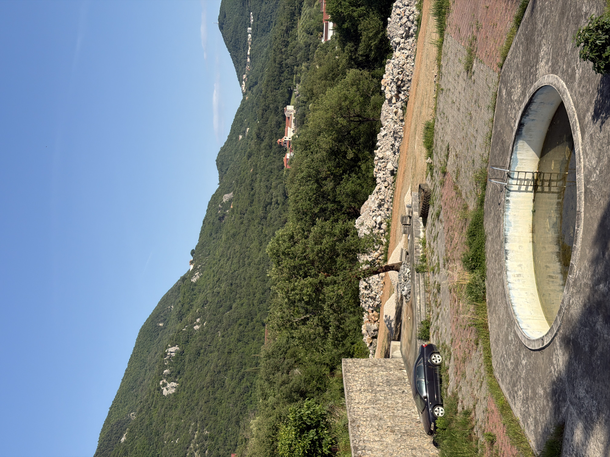 The image depicts an outdoor scene near Bratešići, Montenegro. A steep, forested mountain rises to the left of the frame, with some buildings visible amongst the trees further up. To the right, there's an outdoor concrete circular structure resembling a dry fountain or small pool, with metal railings inside. Adjacent to it, a brick-paved area runs horizontally, bordered by a rough pile of rocks and a retaining wall. A dark-colored car is parked along the bottom edge of the wall. The sky is clear blue.
