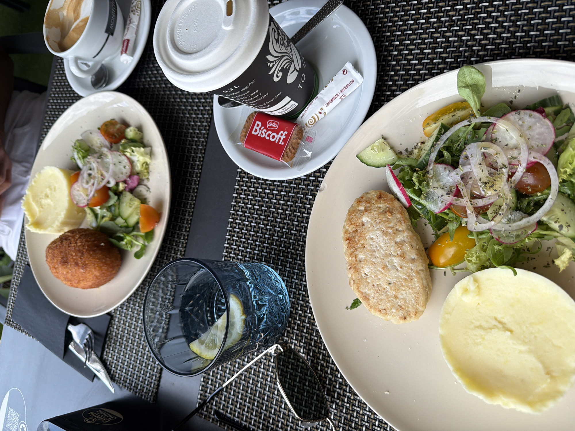 A table set for a meal outdoors. Two plates of food are visible: one with a salad and a breaded patty, and another with a fried ball, mashed potatoes, and a salad. A glass of water with lemon and a pair of sunglasses are on the table, along with a cup of coffee and a small cookie. The table is covered with a woven placemat. The scene appears to be in a restaurant in La Vila Joiosa, Spain.