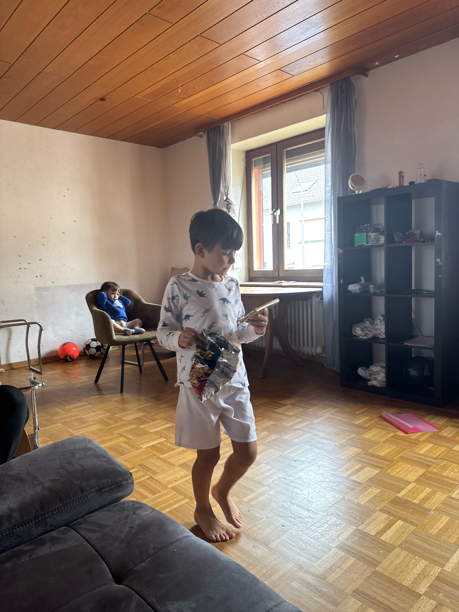 The image shows an indoor scene in a room. A young boy, wearing a white t-shirt with a dinosaur pattern and white shorts, is standing in the foreground, holding a bag of chips and a phone. His feet are bare. In the background, another young boy is sitting in a chair, wearing a blue sweatshirt. Two soccer balls, one red and one black and white, are on the floor near the chair. A dark shelving unit is visible on the right side of the image, filled with various items. A wooden ceiling and wooden floor are prominent features of the room. The scene appears to be a living room or common area. No specific location in Überherrn, Germany, can be determined from the image itself.