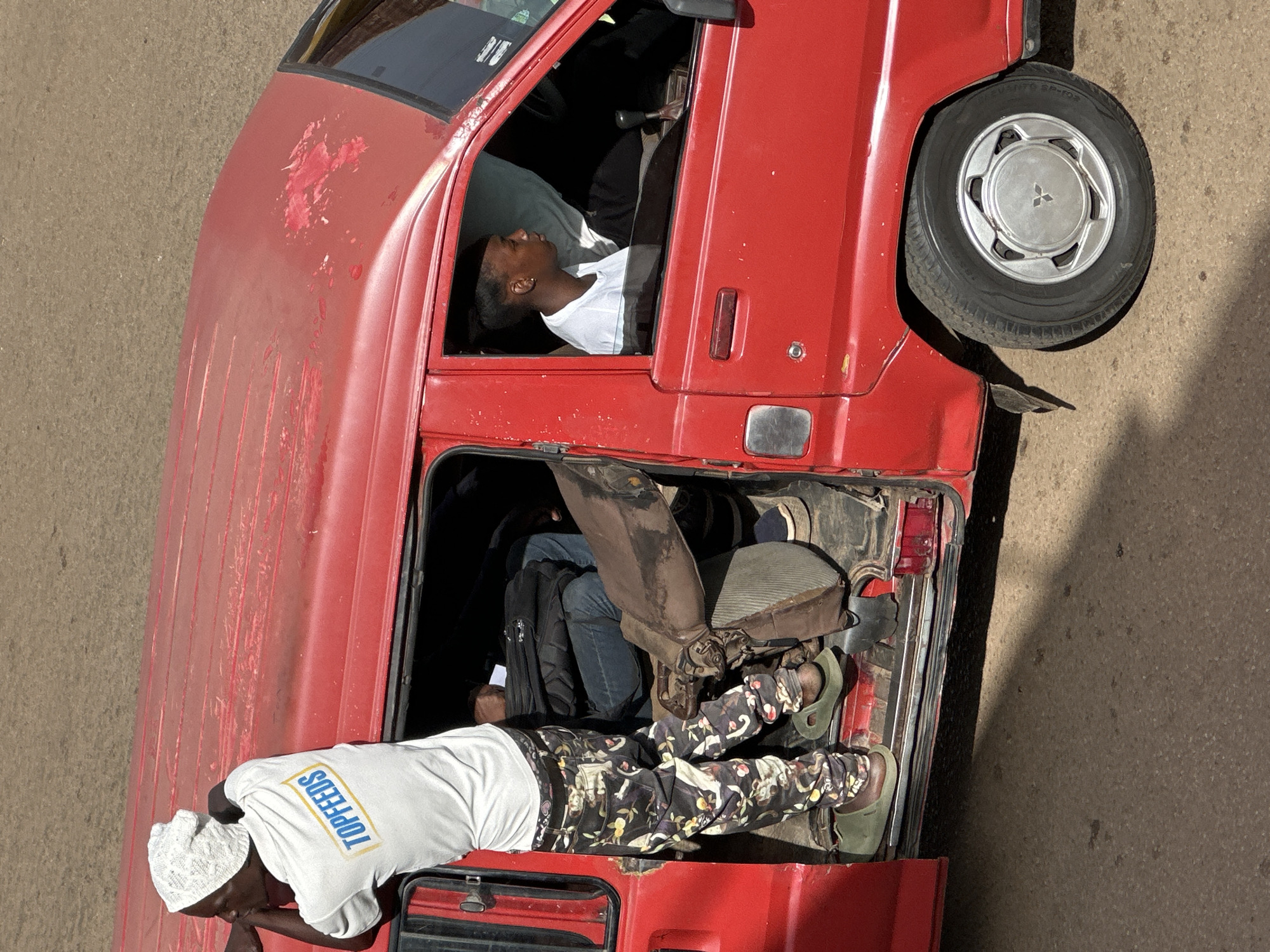 In Bukuru, Nigeria, a bright red, well-worn van is parked on a dirt road. A man, wearing a white t-shirt with "TOPFEEDS" emblazoned on it and a white patterned headscarf, is perched precariously on the edge of the open side door, leaning forward with his right arm resting on the van's roof. He is dressed in patterned camouflage pants and green slip-on shoes. Inside the van, a young man with short hair, wearing a white t-shirt, is seated in the driver's seat, looking out the window. Another person, wearing a dark jacket and blue jeans, is partially visible in the passenger seats. The van's interior is crowded with what appears to be luggage or cargo, including a brown, worn seat or cushion. The van's paint is faded and chipped in several places, revealing patches of primer or rust underneath. The image is captured from a high-angle perspective, casting a shadow on the road.