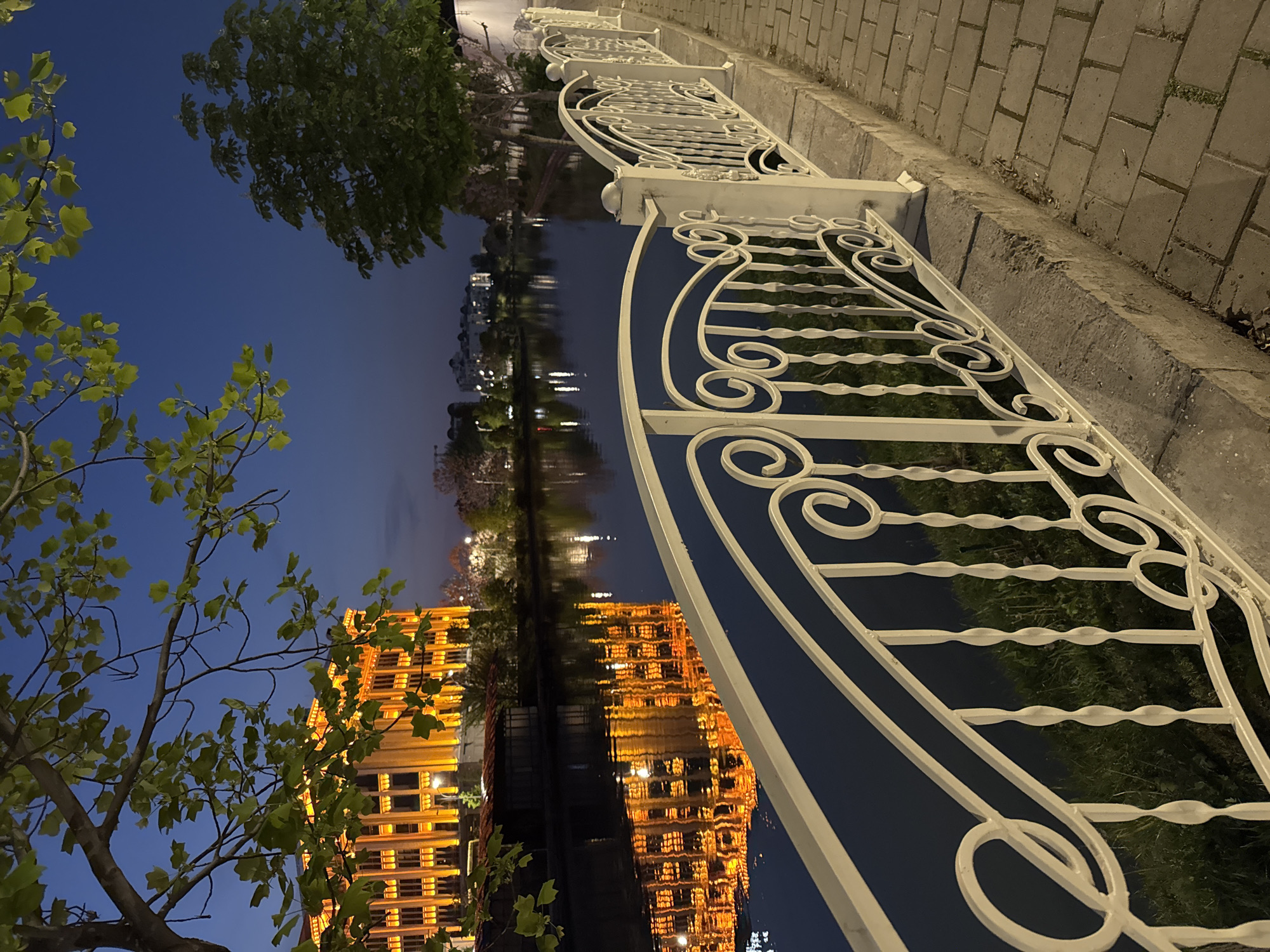 The image shows an ornate, white metal fence running alongside a body of water, reflecting the lit-up facade of a building in Tashkent, Uzbekistan. The building has a warm, orange glow, contrasting with the dark blue sky. A tree with green leaves is visible in the foreground. The fence separates a paved walkway from the water. The photo appears to have been taken at night.