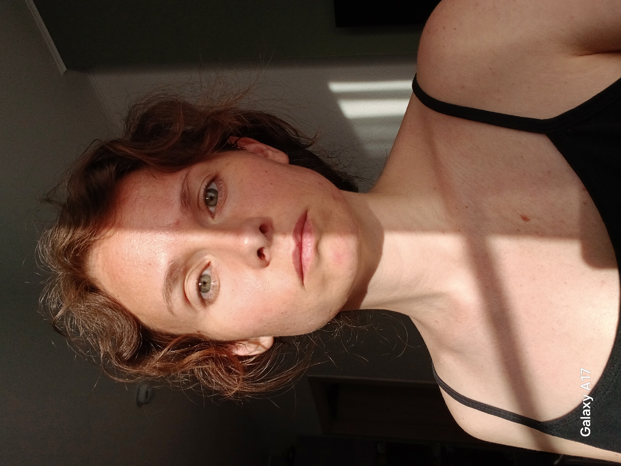A portrait of a woman with light skin and brown curly hair. She is wearing a black tank top. The lighting is dramatic, with stark shadows and bright sunlight creating strong contrast across her face and upper body. The background is dark and out of focus, suggesting an interior space. There are horizontal light stripes on the wall behind her. The camera used to capture the picture might be a Galaxy A17. The image does not provide any information that would suggest that the location is in the city of Yekaterinburg, Russia.