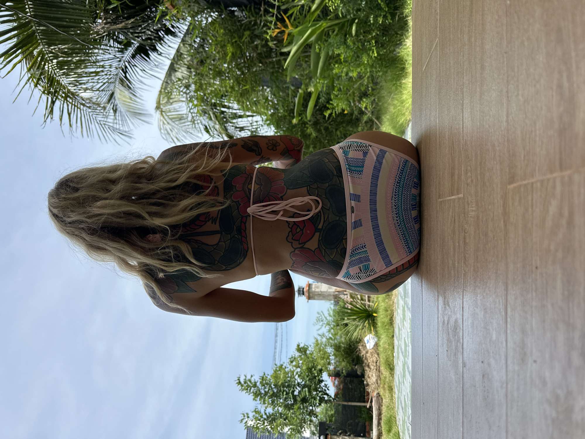 A woman with extensive tattoos covering her back and legs is sitting on a tiled floor, facing away from the camera. She is wearing a pink string bikini bottom with a colorful striped pattern. The background shows lush green vegetation, palm trees, a cloudy sky, and a small lighthouse in the distance, suggesting a tropical coastal location. The setting appears to be an outdoor patio or deck, likely in a resort or private residence in Mũi Né, Vietnam, known for its beaches and resorts.