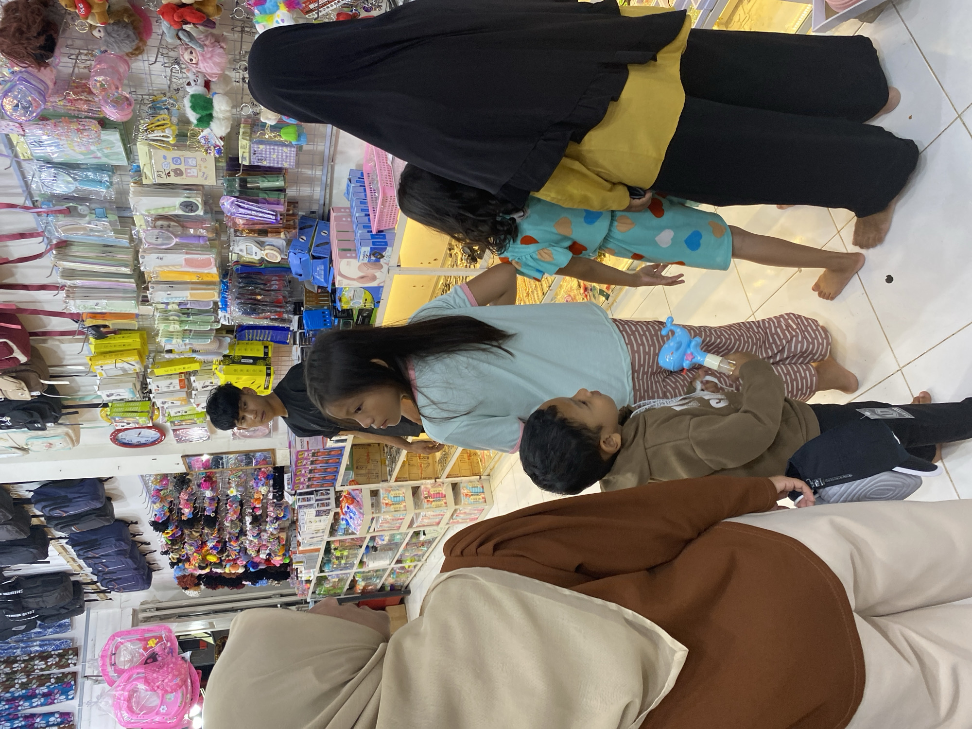 The image depicts an indoor retail setting, possibly a shop, in Lamteumen Barat, Indonesia. The space is densely packed with merchandise displayed on shelves and hanging racks. Various items are visible, including bags, hair accessories, stationery, and trinkets. Several individuals are present in the scene. In the foreground, a person wearing a light brown hijab and a brown top is partially visible, facing away from the camera. To their right, a young boy in a brown hoodie stands, holding a blue toy. Behind him, a young girl in a light blue t-shirt and patterned pants looks to the side. Further to the right, another young girl in a teal patterned outfit is seen from the back. A woman in a black hijab and a yellow top stands at the far right, her back to the camera. The lighting suggests artificial illumination typical of a commercial environment. The floor appears to be tiled. The overall impression is of a bustling retail area filled with a diverse range of consumer goods.