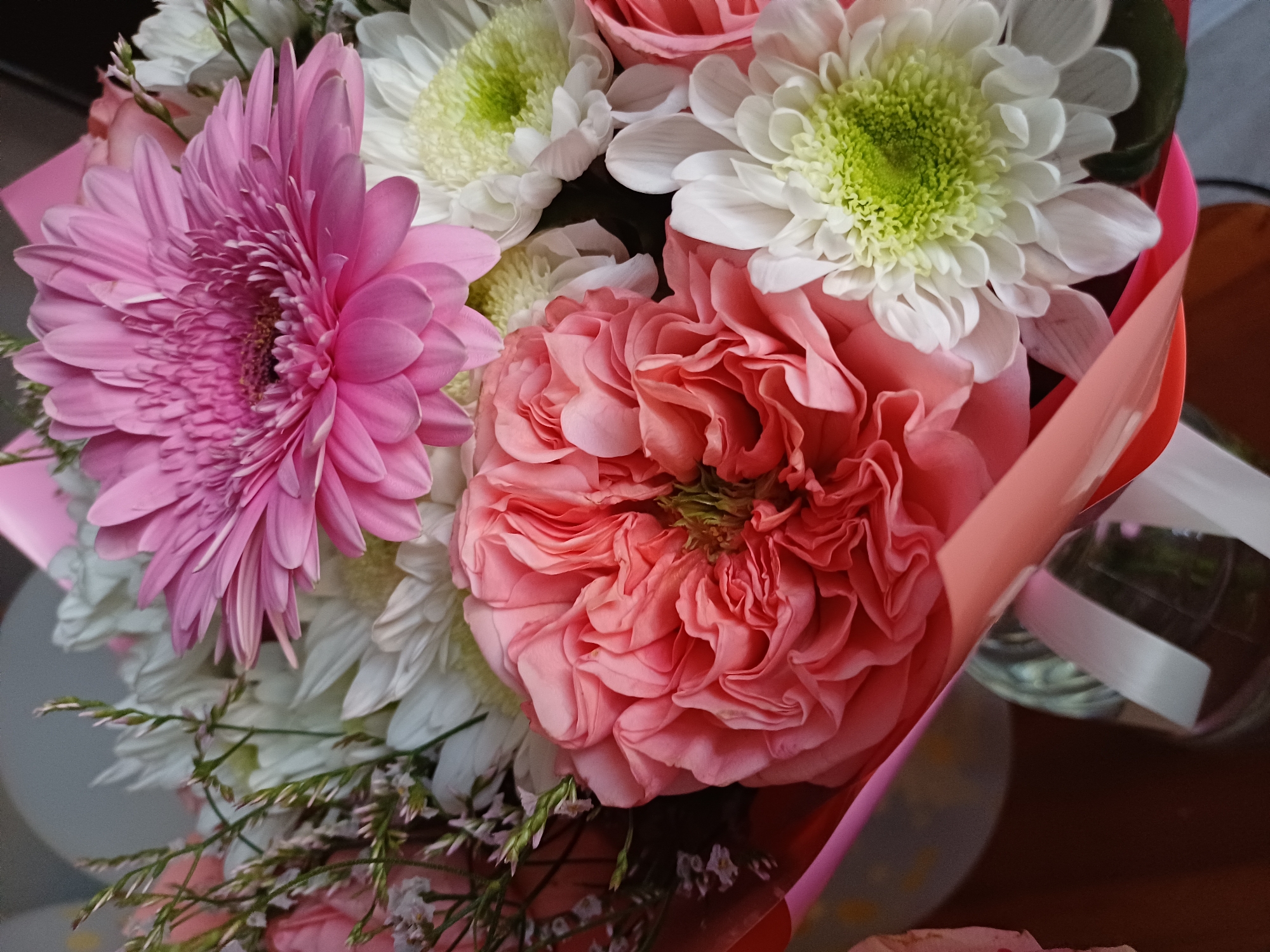 A close-up shot shows a bouquet of flowers in various shades of pink and white. In the foreground, a large, ruffled coral-pink rose is prominently displayed. Behind it, a bright pink gerbera daisy with intricate petals is visible. Interspersed among these larger blooms are smaller white chrysanthemums with green centers and delicate sprigs of greenery and tiny white flowers. The bouquet is partially wrapped in pink and coral-colored paper. The background is blurred, but hints of a glass vase and a brown surface suggest the arrangement is likely indoors. The lighting appears soft, highlighting the textures and colors of the flowers.