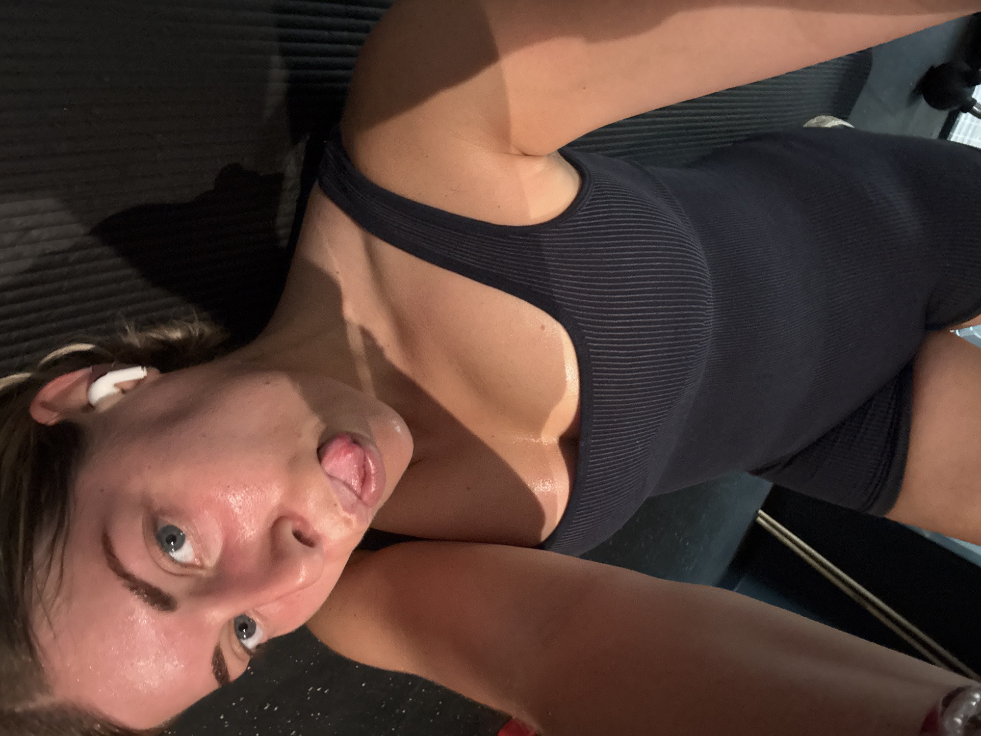 A woman is posing for a selfie. She is wearing a black workout outfit and has an earbud in her left ear. She is sticking her tongue out and looking directly at the camera. Her skin appears to be sweaty. The background is blurry, but it appears she is in a gym, with a black exercise mat visible. The location cannot be determined to be Paris, France, based on the visual information provided.