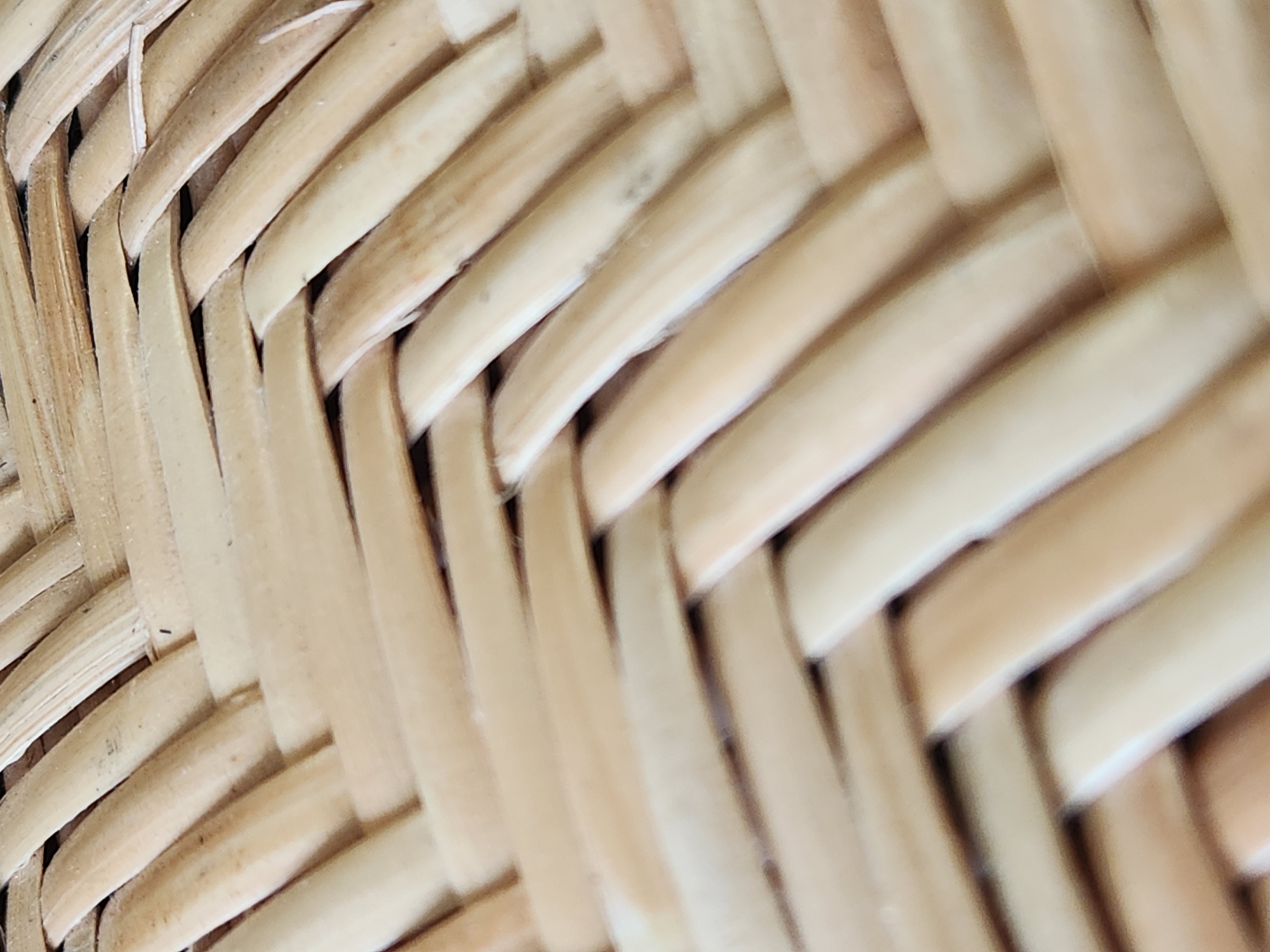 The image shows a close-up, macro shot of a woven material, likely rattan or wicker. The strands are light beige to tan in color and are woven in an over-under pattern. The texture appears slightly rough and organic, with subtle variations in color and some visible imperfections in the weave. The lighting casts soft shadows, highlighting the dimensionality of the material. There are no people, animals, or recognizable landmarks visible. The image is an abstract study of texture and pattern. There is no information that can be used to determine the location in Maitland, United States of America.