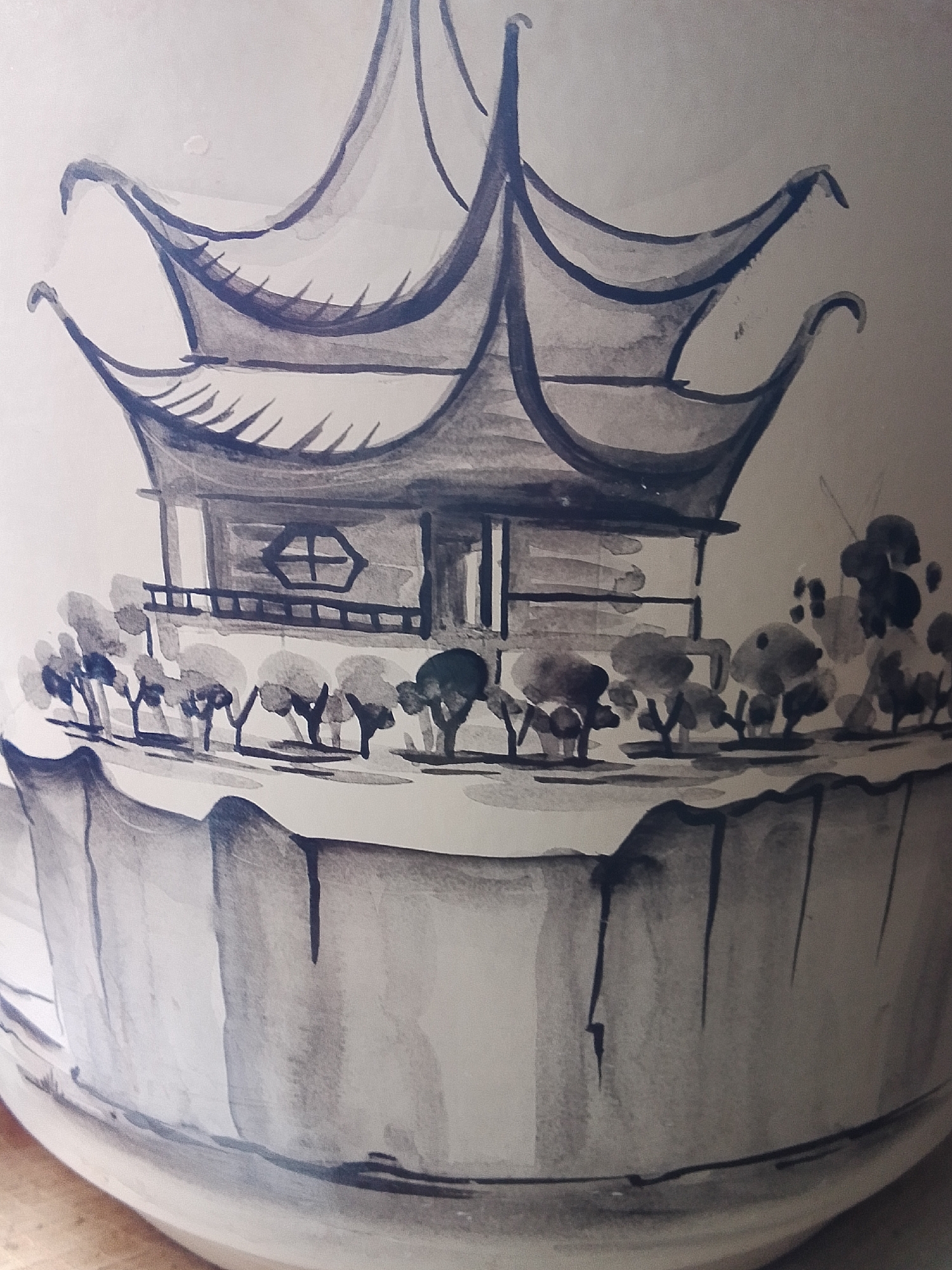 The image shows a close-up of a painted object, possibly a vase or pot. The painting depicts a stylized scene of a pagoda-like structure with multiple tiered roofs. A small balcony with a railing is visible beneath the main roof. In front of the structure, there's a line of small trees or bushes. The painting is done in a blue-gray ink wash style. The lower portion of the object is also painted with vertical strokes that suggest a base or foundation. There are no people or other obvious figures in the scene. The background is a plain white or light color, suggesting the surface of the object. The overall style is reminiscent of traditional East Asian art. The city of 1-Go Maya, Russia, is not depicted nor is there any information in the image to support this claim.