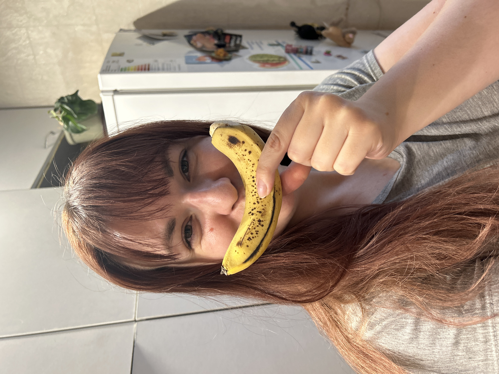 A woman with red-brown hair and bangs is holding a ripe banana up to her face, the banana's curve mimicking a smile. She is wearing a gray shirt. The scene appears to be indoors, possibly in a kitchen, given the presence of a white refrigerator in the background. The refrigerator has magnets or decorations on its surface. The background also includes a potted plant on a counter near a white tile wall. The image is oriented with the woman and banana rotated counter-clockwise. There is no information in the image to confirm that the location is in the city of Фоки, Russia.