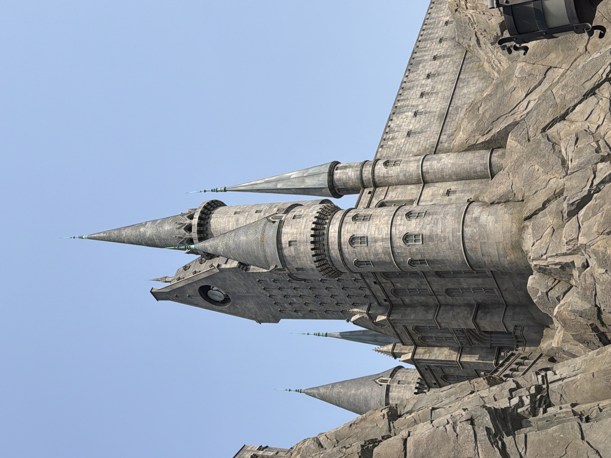 The image depicts a section of Hogwarts Castle, a themed structure within a theme park. The castle is constructed of grey stone and features turrets, spires, and various architectural details that give it a medieval, fantastical appearance. It is built on a rocky, mountainous base, which blends with the castle's design. The background consists of a clear, light blue sky. The exact location within Tongzhou District, China, cannot be identified from the provided image.