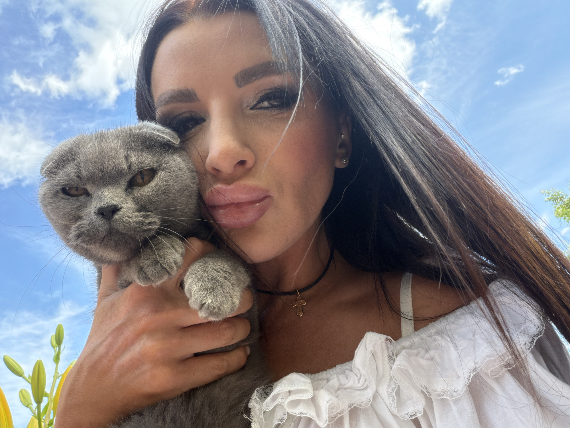 The media file is a selfie of a woman holding a Scottish Fold cat. The woman has long, dark hair, full lips, and is wearing makeup. She has a gold cross necklace and a white ruffled off-the-shoulder top. The cat is grey and appears to be held closely to the woman's face. The background shows a bright blue sky with scattered white clouds, suggesting it is daytime and fair weather. Yellow flower buds are visible in the lower left corner. The scene appears to be outdoors, possibly in a garden or natural setting. The woman's expression, with her lips pursed, and the cat's slightly worried look, suggest a playful or affectionate moment. There is no visible text in the media file. The location context is Novaya Usman', Russia.