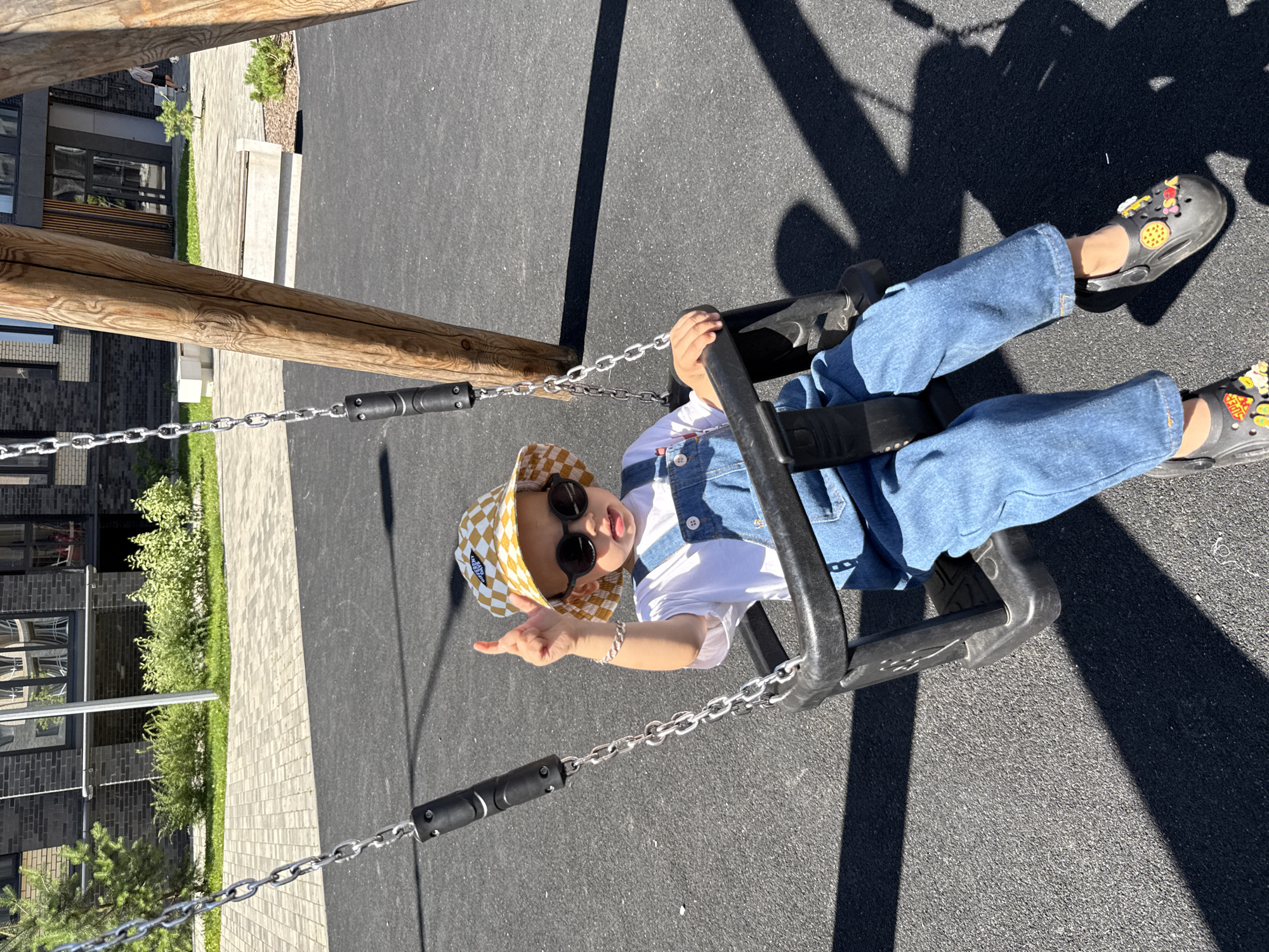 A young child is seated on a black swing with a white shirt and blue denim overalls. The child wears a yellow and white checkered bucket hat and round black sunglasses. Their right index finger is raised. The swing is suspended by metal chains from a wooden structure. The ground beneath the swing is black asphalt, with dark shadows cast by the wooden supports. In the background, a modern brick building with multiple windows is visible, surrounded by green bushes and a paved walkway. The location is identified as Rusanovo, Russia.