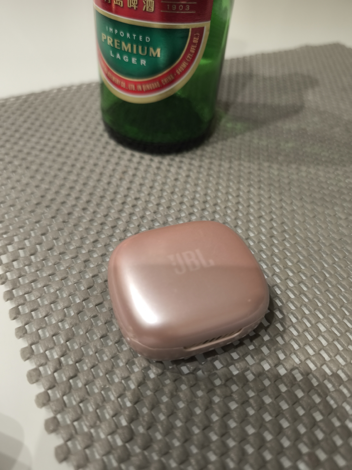 The image shows a pink JBL earbud case on a gray woven placemat next to a green beer bottle. The scene is indoors, with the placemat appearing to be on a table.