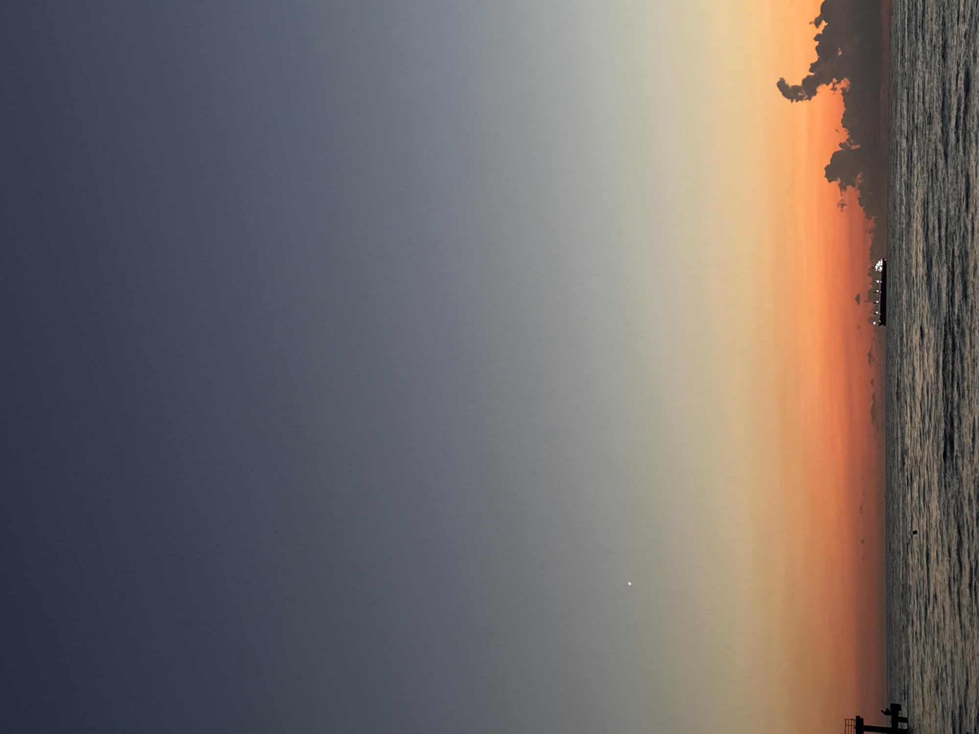 The image depicts a seascape at sunset, with the sky transitioning from a deep blue at the top to vibrant oranges and reds near the horizon. A small, bright point, possibly a star or planet, is visible in the upper left quadrant of the sky. A large cargo ship is sailing on the water in the middle ground, illuminated by its own lights. To the right of the ship, dark, stylized clouds are present. In the lower left of the image, a pier structure is visible with two silhouetted figures on it. The water is rippled, indicating a moderate sea state. This scene is consistent with a view of the Black Sea coast in Batumi, Georgia, known for its sunset vistas.
