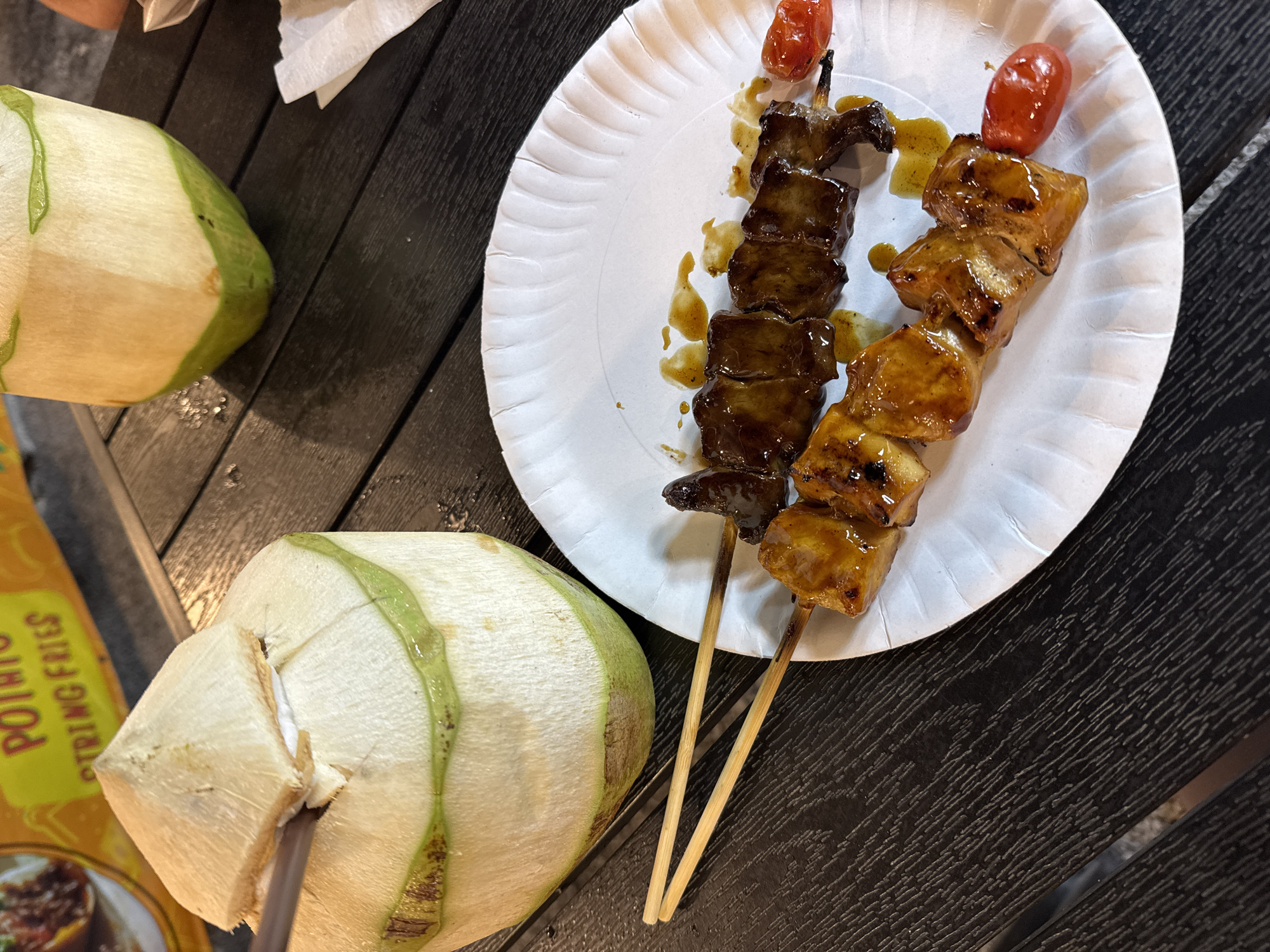The image captures a casual food scene, likely on a street food vendor's table in Baan Bo Phut, Thailand. Two young coconuts sit on a dark wooden surface, next to a white paper plate holding two skewers of grilled meat. One skewer has dark, possibly beef, cubes, while the other has lighter, glazed chunks, possibly chicken or pork. Each skewer is topped with a grilled cherry tomato. A yellow advertisement for "potato string fries" is partially visible.