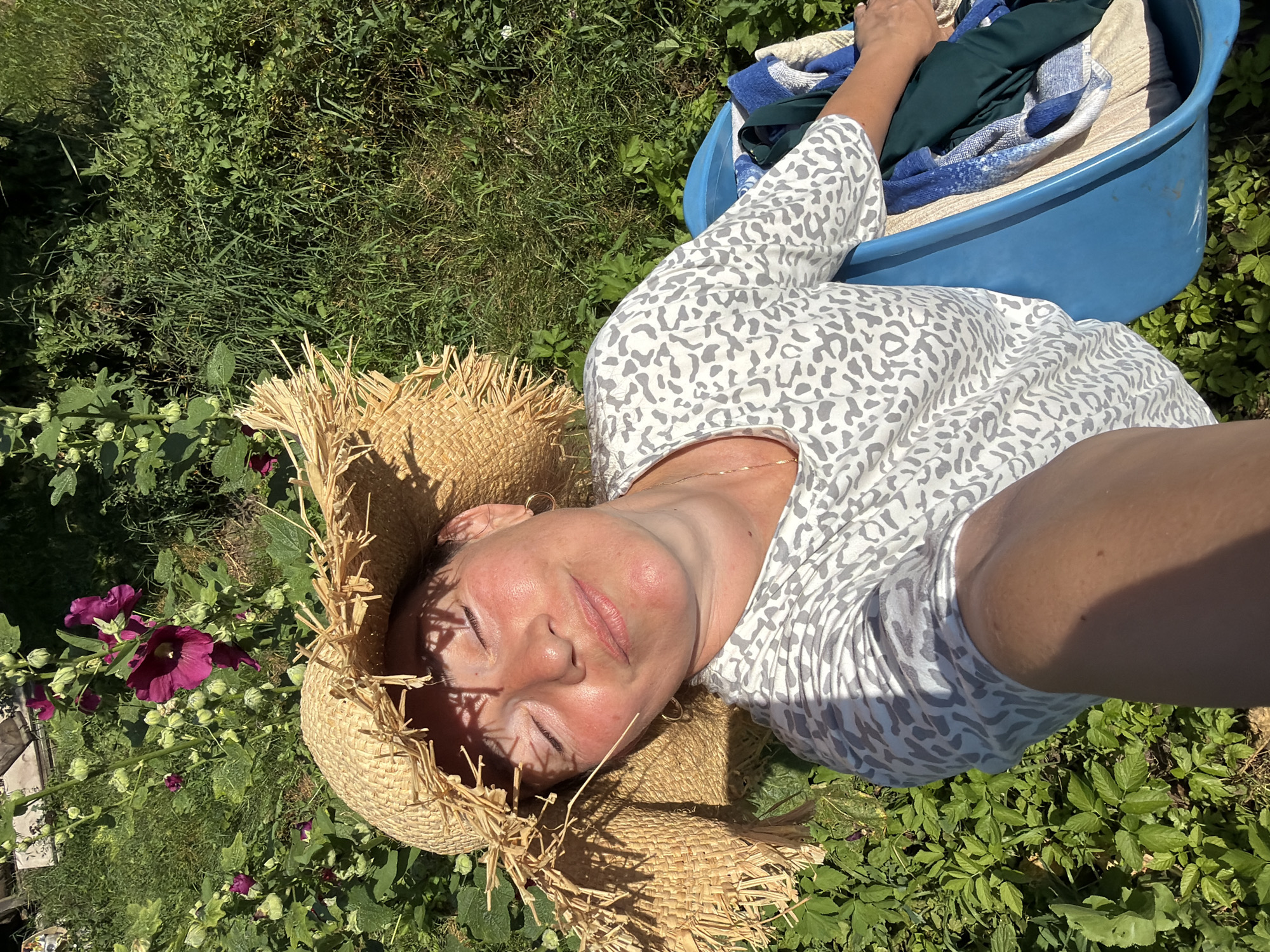 A woman wearing a straw hat and a grey and white patterned t-shirt lies in a blue bin outdoors, with her eyes closed and a peaceful expression on her face. Sunlight casts shadows across her face from the hat. She is surrounded by lush green foliage and some purple flowers. In the bin with her are some folded items of clothing, including blue and white towels and a dark green garment. The scene appears to be in a garden setting, possibly in the city of Olkhovka, Russia, given the mention of the location.
