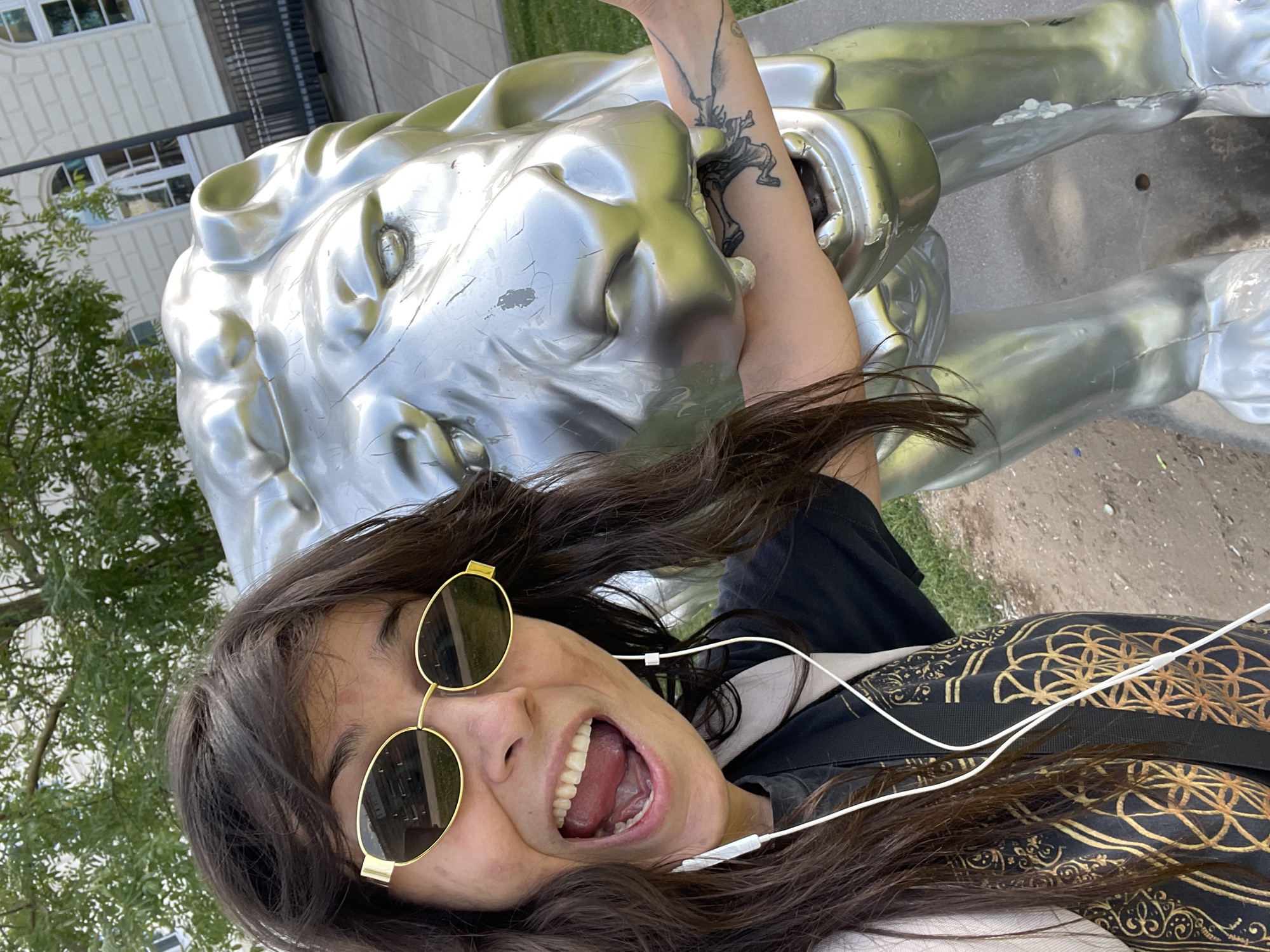 An outdoor, eye-level, medium shot shows a woman with long, dark hair, yellow oval sunglasses, and white earbuds, taking a selfie next to a large, silver lion statue. The woman is smiling with her mouth open, and her right arm is positioned as if being bitten by the statue's mouth. She is wearing a black t-shirt with a gold geometric pattern and a light-colored backpack. The lion statue is made of a reflective material and shows some wear and tear, with visible scratches and peeling paint. The background includes green trees, a building with white facade and arched windows, and a paved area. The scene appears to be a public space, possibly a park or plaza in Wuppertal, Germany. The lighting suggests daytime.
