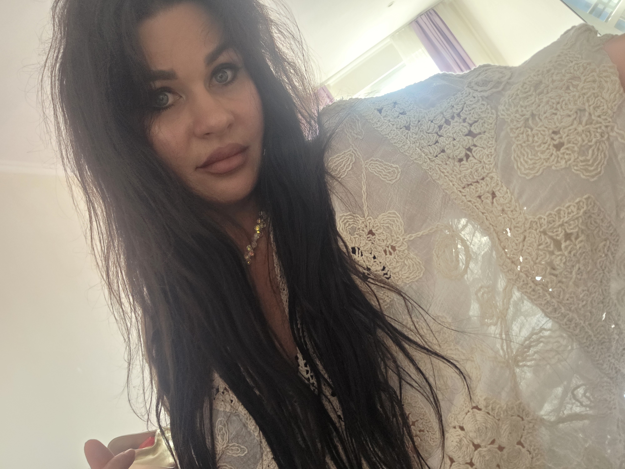 A low-angle, indoor selfie shows a woman with long, dark hair, wearing a white, crochet-detailed top. She is looking at the camera, with her head tilted and her gaze directed slightly to the right. Her eyes are a striking blue. The background is out of focus and appears to be a room with natural light, possibly from a window with purple curtains visible on the right side. The overall lighting suggests daytime. There are no discernible landmarks or contextual clues to suggest the location is Sochi, Russia.