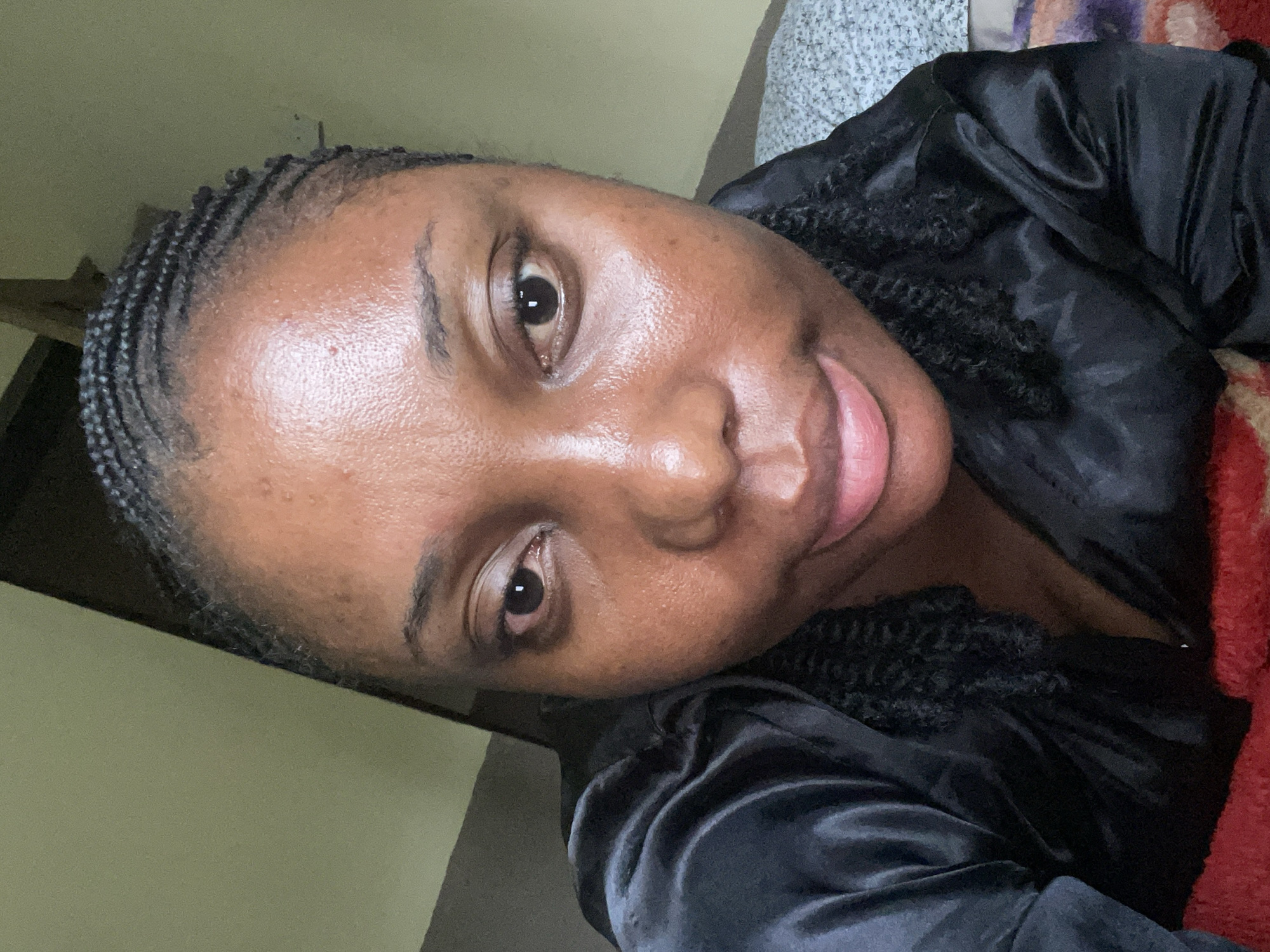 A close-up, indoor shot focuses on a Black woman with her head tilted. She is wearing a black, silky garment, possibly a robe or pajama top. Her hair is styled in small braids that go straight back from her forehead, with textured hair visible near her cheek. Her skin appears shiny. The background includes a light green wall and an out-of-focus bed with a patterned sheet. It is not possible to determine the location as the city of Jos, Nigeria from the image. There is no indication of activity.