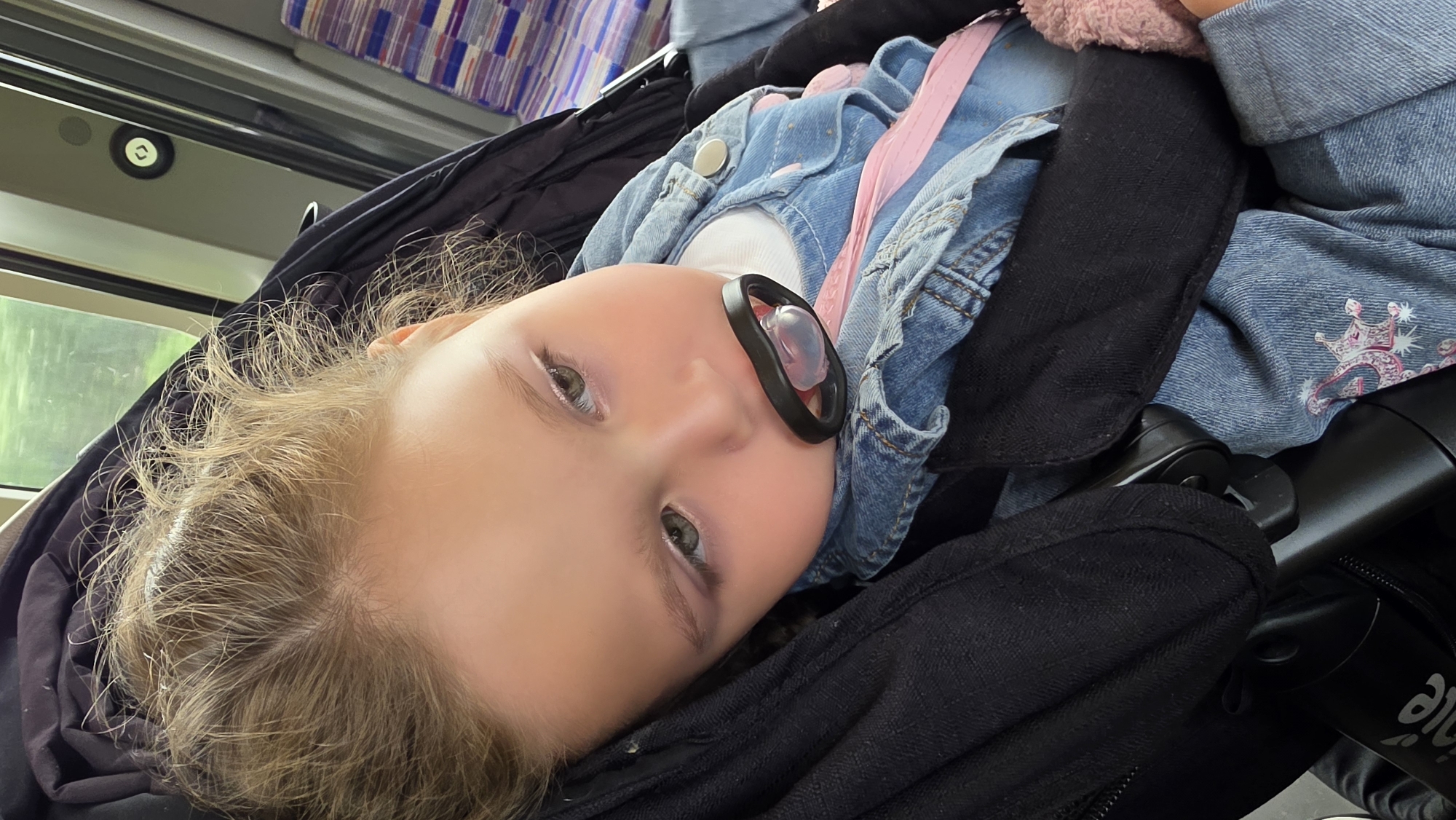 A young child lies in a stroller, likely on a train in London, United Kingdom. The child has fair skin, blonde curly hair, and is wearing a denim jacket with a pink strap and a pacifier in their mouth. Part of the stroller and the interior of the train are visible in the background. The train's window, a colorful, upholstered seat, and the upper portion of the train interior are discernible.
