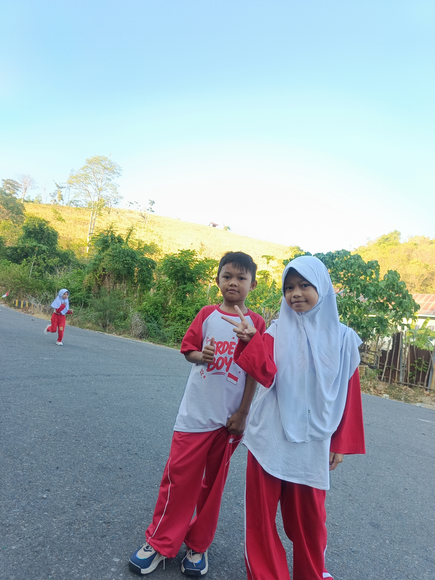 Two children, a boy and a girl, pose for a photo on a paved road, likely in Ilotidea, Indonesia, with a hillside covered in vegetation in the background. The boy, on the left, wears a white t-shirt with red sleeves and red pants, and he makes a peace sign with his hand. The girl, on the right, wears a white hijab and a red outfit with white accents, and she is also facing the camera. Another child, wearing a similar red and white outfit, runs on the left side of the road in the distance. The sky is clear and blue.
