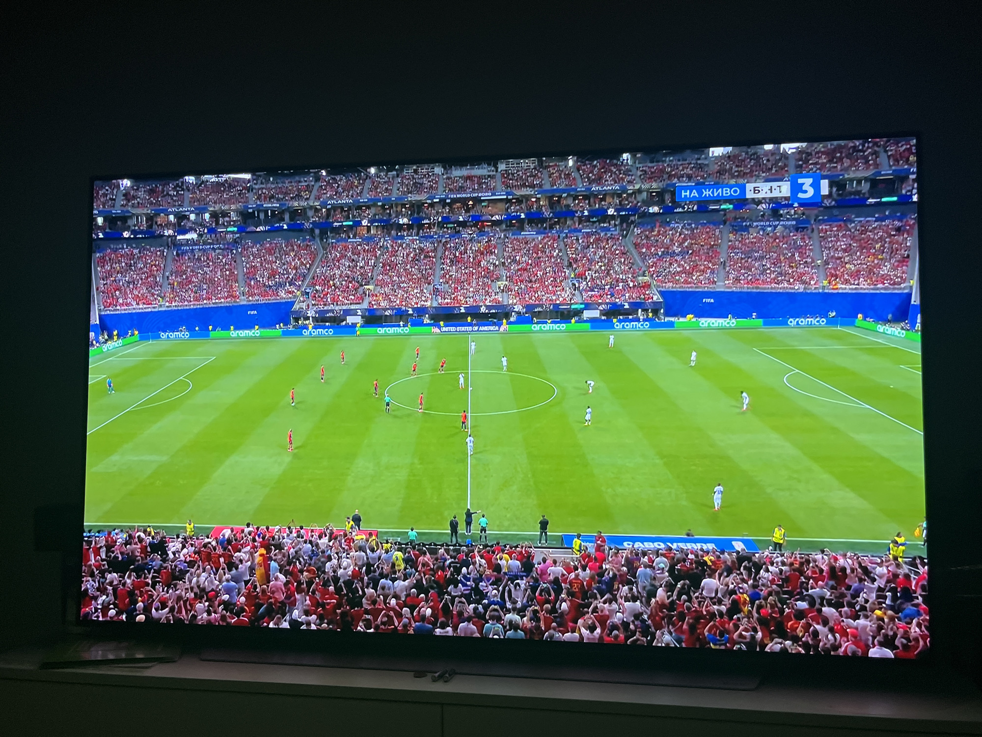 The image shows a wide-angle view of a soccer match being played in a stadium filled with spectators. The field is green with white markings, and the stadium seating is packed with people wearing red and white clothing. There is a large scoreboard visible in the upper right corner of the screen, displaying text in Bulgarian and a score of "3-1". The text "НА ЖИВО" is visible on the scoreboard, which translates to "LIVE". The stadium is identified as being located in the city of Varna, Bulgaria. The event is branded as "FIFA WORLD CUP 2026 QUALIFIERS". The overall scene suggests a vibrant and energetic sports event.