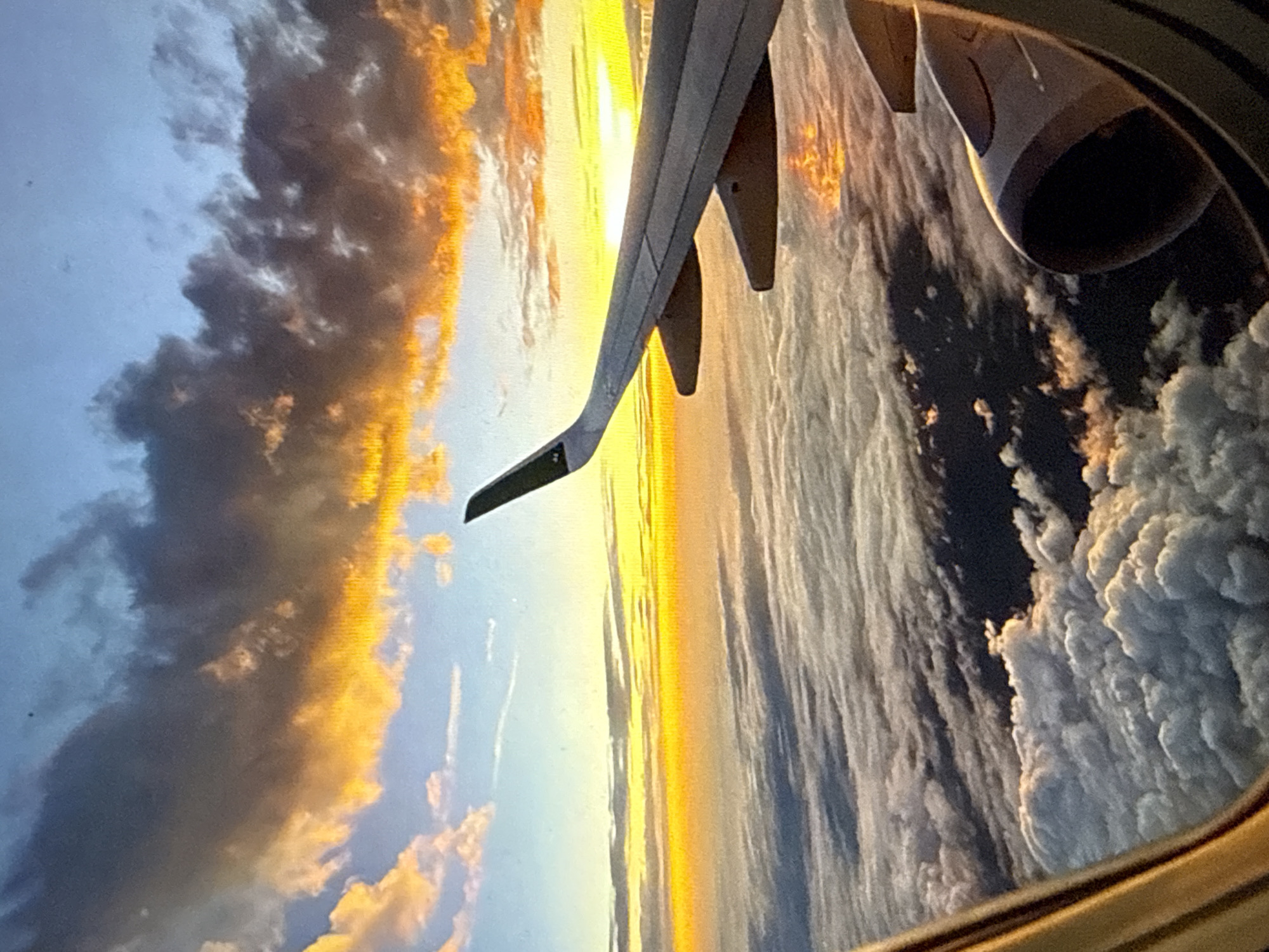 An aerial view from an airplane window, taken during a flight over the general vicinity of Kardzhali, Bulgaria. The image depicts a dramatic sunset or sunrise, with the horizon dominated by intense golden-yellow light. In the foreground, the lower-left shows the curved, reflective frame of the airplane's window. The mid-right displays a portion of a light grey aircraft wing with visible flap track fairings and a dark-tipped winglet. Below the wing, a partial view of a dark, circular engine nacelle is present. Below the aircraft, a dense, undulating layer of white cumulus clouds stretches to the horizon, reflecting the golden light. Above the wing, the sky transitions from vibrant orange and yellow near the horizon to light blue overhead, interspersed with dark, textured clouds featuring bright golden edges. No people or specific topographical features of Kardzhali are visible within the frame, which concentrates on the atmospheric conditions and aircraft components.
