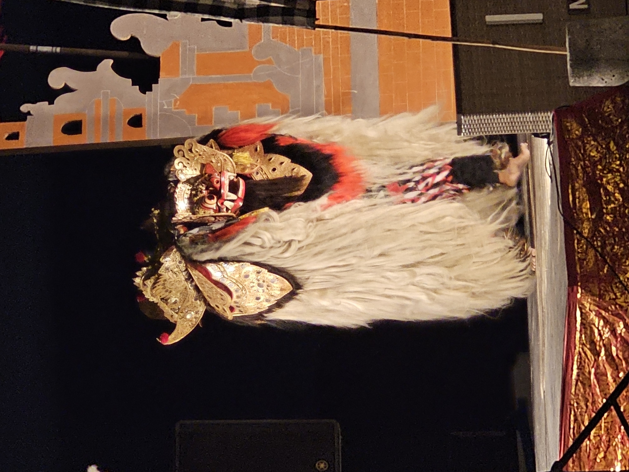 Captured at night, the image reveals a Barong landung performance in Denpasar, Indonesia. The focus is on the Barong landung effigy, a large, white-haired, ornamental mask with a red face and golden adornments. The Barong landung is positioned in front of a backdrop featuring traditional Balinese architectural elements. A red and gold fabric drape adds to the ornate setting. The dark background suggests an outdoor stage or public space lit for the performance.