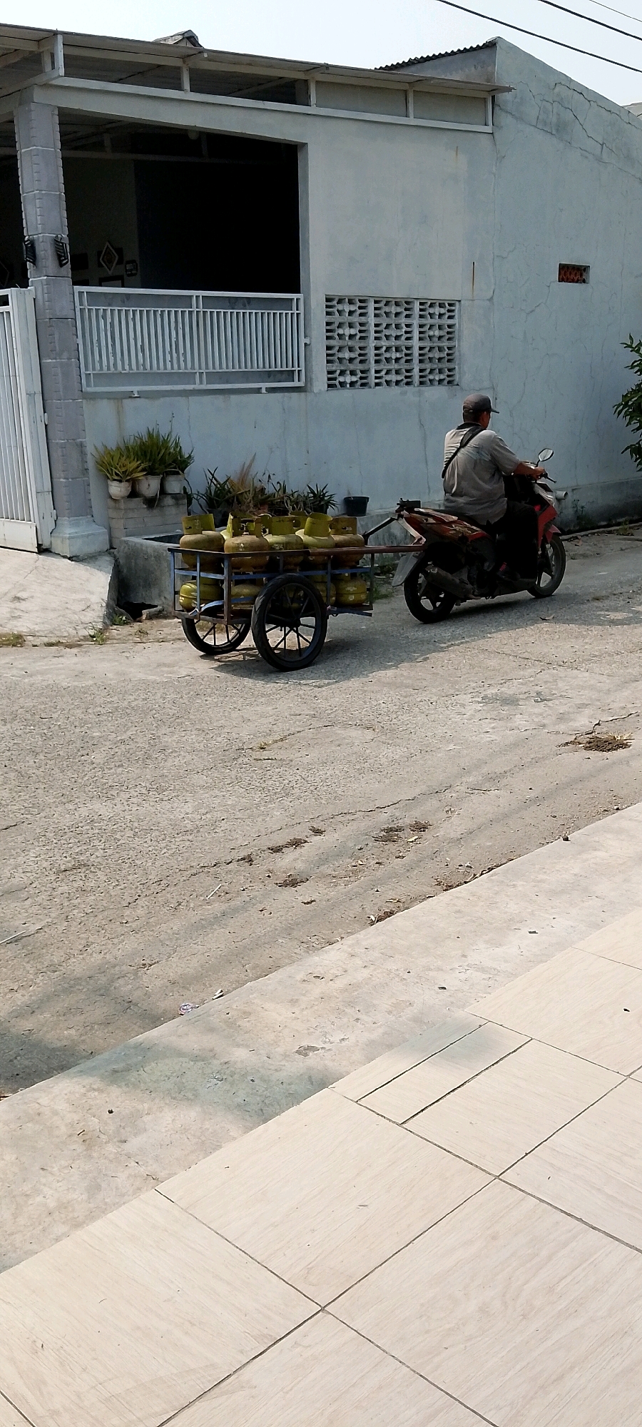 In Satriamekar, Indonesia, a man wearing a grey shirt and a cap rides a red and black motorcycle. He is pulling a two-wheeled cart filled with multiple yellow LPG gas cylinders. The cart is made of metal with wooden planks. The motorcycle and cart are on a paved road in front of a light-colored building with white railings and decorative vents. Several potted plants are visible near the building. The scene appears to be outdoors during the day.