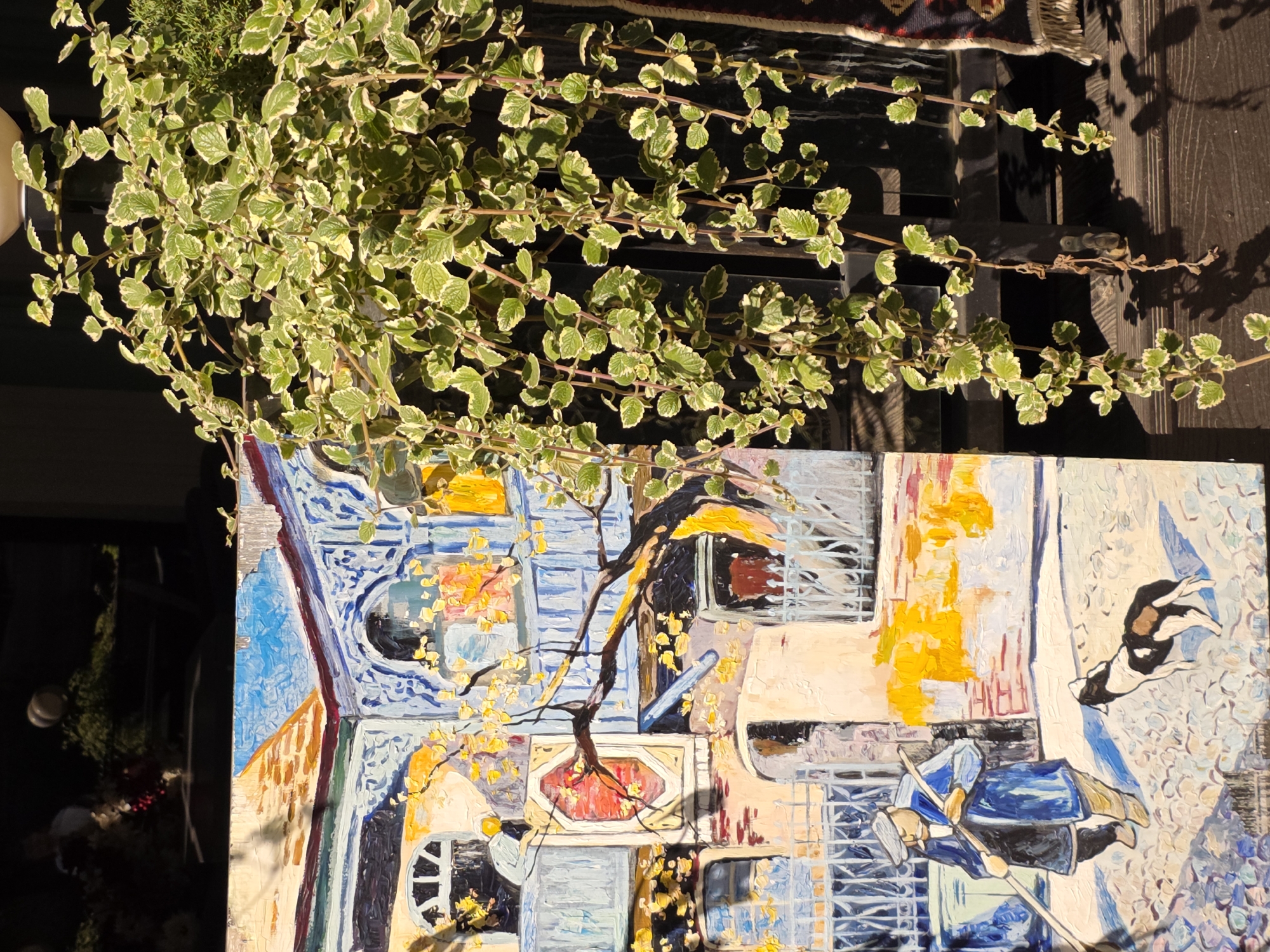 This is a photograph of a painting in Rostov-on-Don, Russia, displayed outdoors. The painting depicts a scene with a man in a blue coat and cap pushing a broom on a cobblestone street, accompanied by a black and white dog. In the background, there is a building with blue and yellow architectural details, balconies, and windows. A tree with yellow leaves extends into the frame from the left. To the right of the painting, a trailing houseplant with green and white variegated leaves hangs down, partially obscuring a dark wooden surface and a patterned rug with fringe. The lighting suggests it is a sunny day, casting shadows.