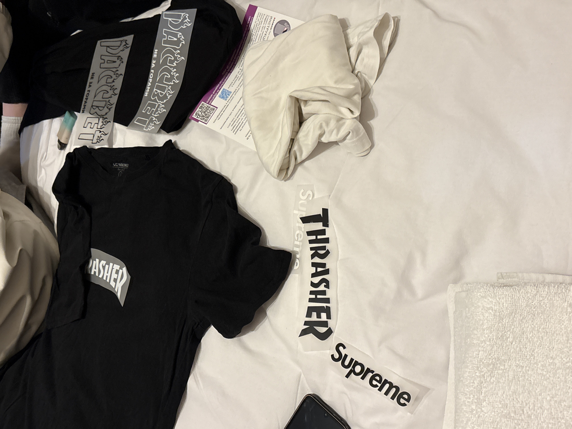 The image depicts a scene on a white bedsheet, with various clothing items and stickers scattered across it. A black t-shirt with a white "Thrasher" logo is positioned on the left side of the image. To its right are two stickers, one reading "Thrasher" vertically and the other "Supreme" horizontally. Two black t-shirts with Cyrillic text, "Packet" (Пакет) and "Not Behind the Mountains" (Не за Горами) stickers are present near the upper-left corner. A crumpled white t-shirt rests above the "Thrasher" shirt. There is a pamphlet written in Russian and a white towel in the lower right. A black smartphone is positioned near the bottom. There is no information regarding the location in the city of Yerevan, Armenia.