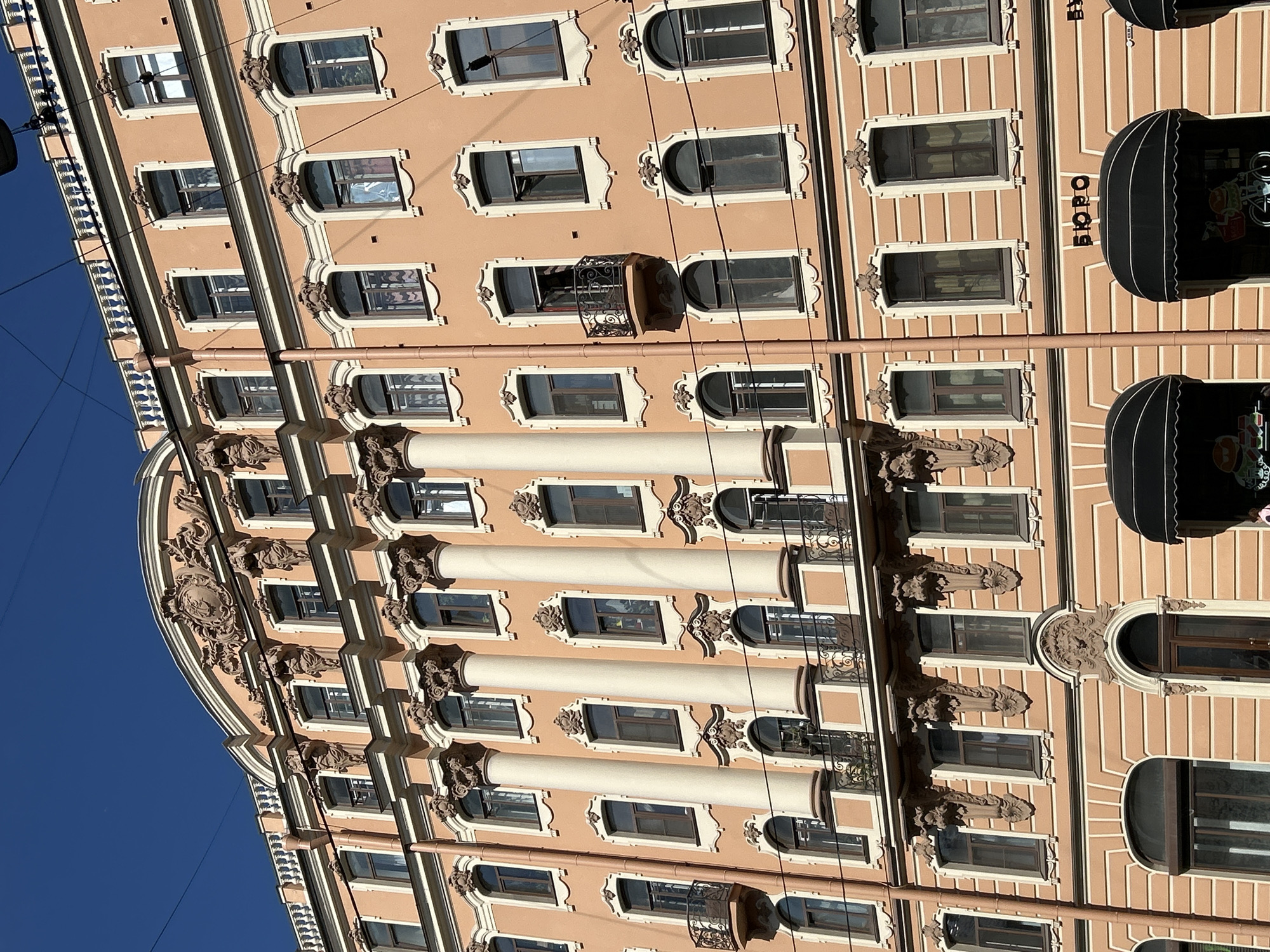 A low-angle view captures the architectural facade of a building in Saint Petersburg, Russia. The structure exhibits a salmon-colored exterior, adorned with rows of windows that feature ornate, creamy-white trim. The building's facade is punctuated by vertical columns that run the height of multiple stories, adding a sense of grandeur and classical design. Small wrought-iron balconies are occasionally present, projecting from some of the windows. On the right side of the building, black awnings extend over the storefronts, marked with the word "Bopo" written vertically. Electrical lines cut across the clear blue sky that forms the backdrop, and the building curves on the left side, reflecting a portion of the elaborate decorations near the corner.