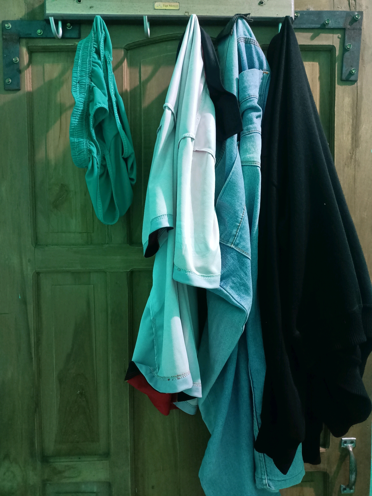 An overhead shot shows several pieces of clothing hanging on hooks attached to a wooden door frame in what appears to be a room in Ngargosari, Indonesia. From left to right, there is a pair of teal-colored shorts, a light blue long-sleeved shirt, a black t-shirt, and a denim jacket. The clothing is casually arranged, suggesting it is being air-dried or stored. The wooden door has a rustic appearance with visible grain and panels. The lighting in the room is dim and casts shadows, creating a slightly moody atmosphere.