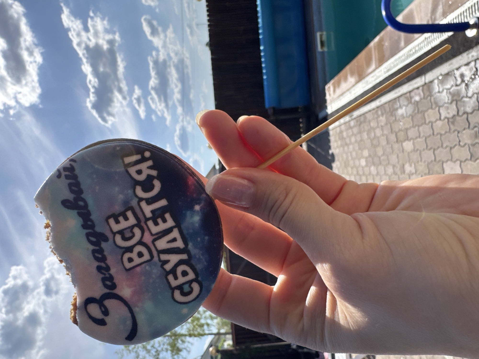 A person's hand holds a round cookie on a stick against a backdrop of a partly cloudy sky. The cookie has a bite taken out of it, and it is decorated with an image of a galaxy and the Russian text "Загадывай - все сбудется!", which translates to "Make a wish - everything will come true!". In the background, there are structures suggesting an outdoor recreation area, including a blue water slide and a brick-paved area. The location is Chaikovsky, Russia.