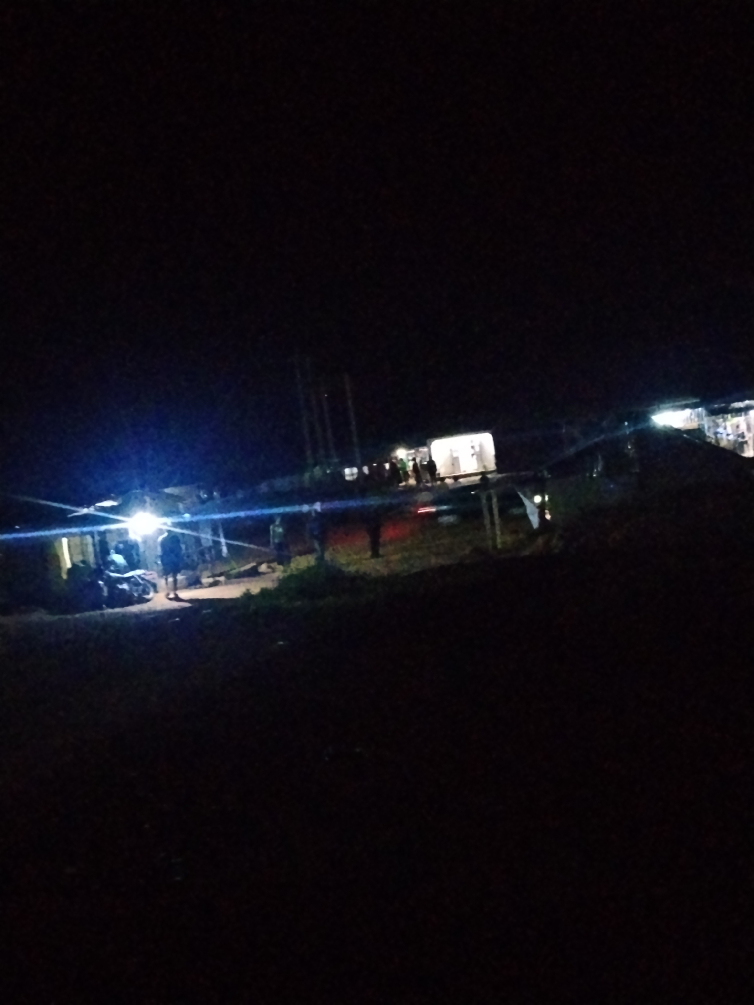 This is a nighttime image taken outdoors in Durumi, Nigeria. The scene appears to be a gathering or event taking place in a somewhat rural or undeveloped area, indicated by the presence of unpaved ground and vegetation. Several people are visible, most of them standing or walking. In the foreground, there is a motorcycle, and a person is standing near it.
The setting includes some structures that look like stalls or small shops, illuminated by bright artificial lights, creating a stark contrast with the surrounding darkness. These lights also cause lens flares. A few vehicles are also present, including what appears to be a white van or truck in the background, and possibly a car. The overall impression is of a lively social gathering, perhaps a market or a local event happening after dark. The time of day is clearly night, and the weather is not discernible from the image. There is no visible text in the image. The emotions of the individuals are not clear due to the low light conditions and image quality.