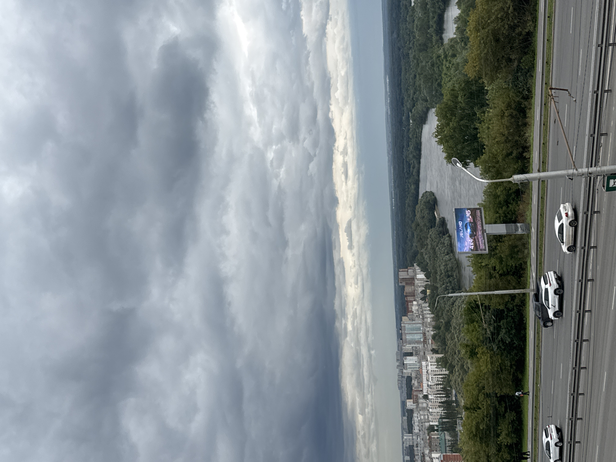 The image captures a panoramic view of the city of Ufa, Russia, likely from an elevated vantage point overlooking the Belaya River. The foreground features a multi-lane highway with several white cars and a black SUV driving. A street light stands prominently on the right side of the highway. In the middle ground, a dense forest borders the river, which curves through the landscape. On the left, a portion of the Ufa cityscape is visible, characterized by numerous apartment buildings of varying heights and architectural styles. A large billboard advertising "JELAND 4x4 J6" is positioned amidst the trees. The sky is overcast with heavy, dark clouds, suggesting inclement weather. Two figures are visible walking on a path to the left of the highway, near the edge of the forested area.