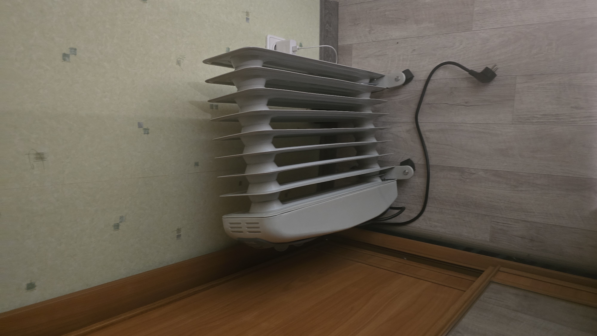 The image shows a white electric oil-filled radiator placed on a light wood-grain laminate floor next to a wooden cabinet or door. The radiator has multiple fins and a control panel on the side. A black power cord extends from the radiator to the right. In the background, the wall is covered with a light-colored wallpaper with a subtle pattern. A white electrical outlet is visible on the wall behind the radiator. The scene appears to be indoors, likely in a residential setting. There are no people or animals present. The lighting suggests it is daytime. The location is identified as Ukhta, Russia.