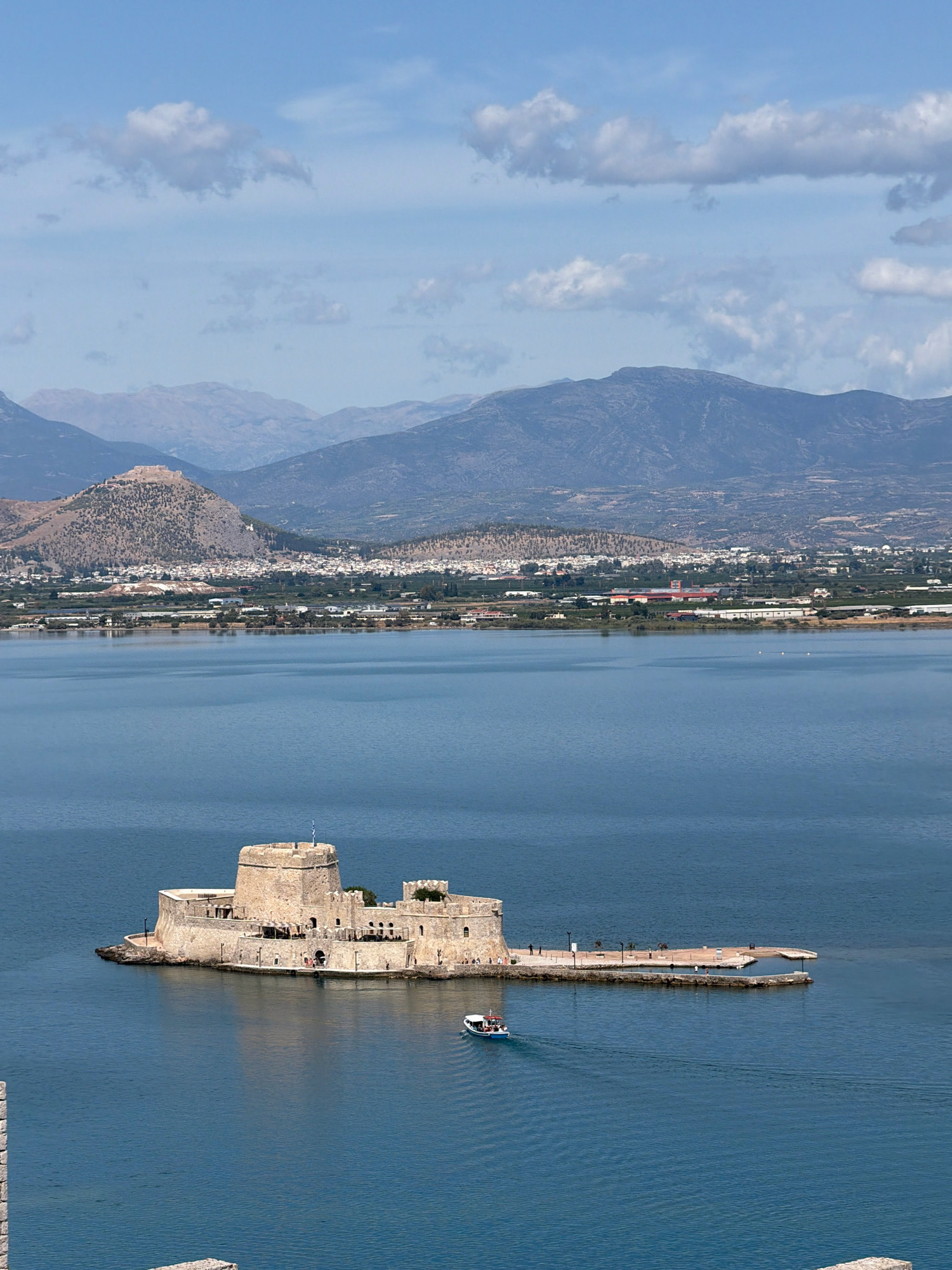 A castle sits on a small island in the middle of a body of water. The castle is made of stone and has a flag flying from its highest tower. A small boat is sailing on the water in front of the castle. In the background, there are mountains and a town. The sky is blue with some clouds. The location appears to be the Bourtzi fortress in Nafplio, Greece.