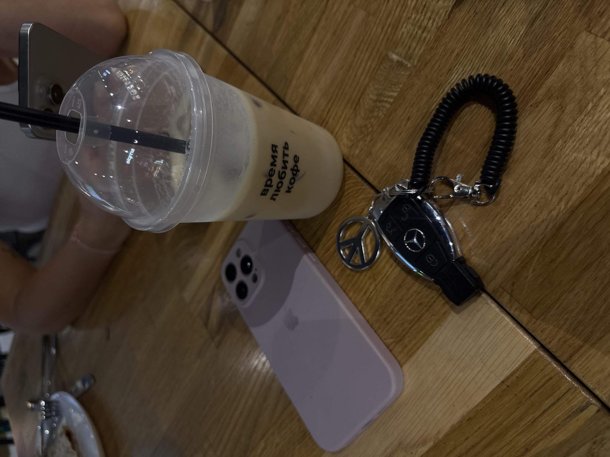 On a wooden table, there's a clear plastic cup filled with iced coffee; the text "время любить кофе" (time to love coffee) is printed on it. Beside it sits a light purple iPhone and car keys attached to a coiled black keychain and a peace symbol charm. The scene appears to be a café or restaurant in Moscow, Russia, based on the Cyrillic text. A glimpse of a person's arm and smartphone is visible near the upper left corner, and part of a plate with food is on the lower left.