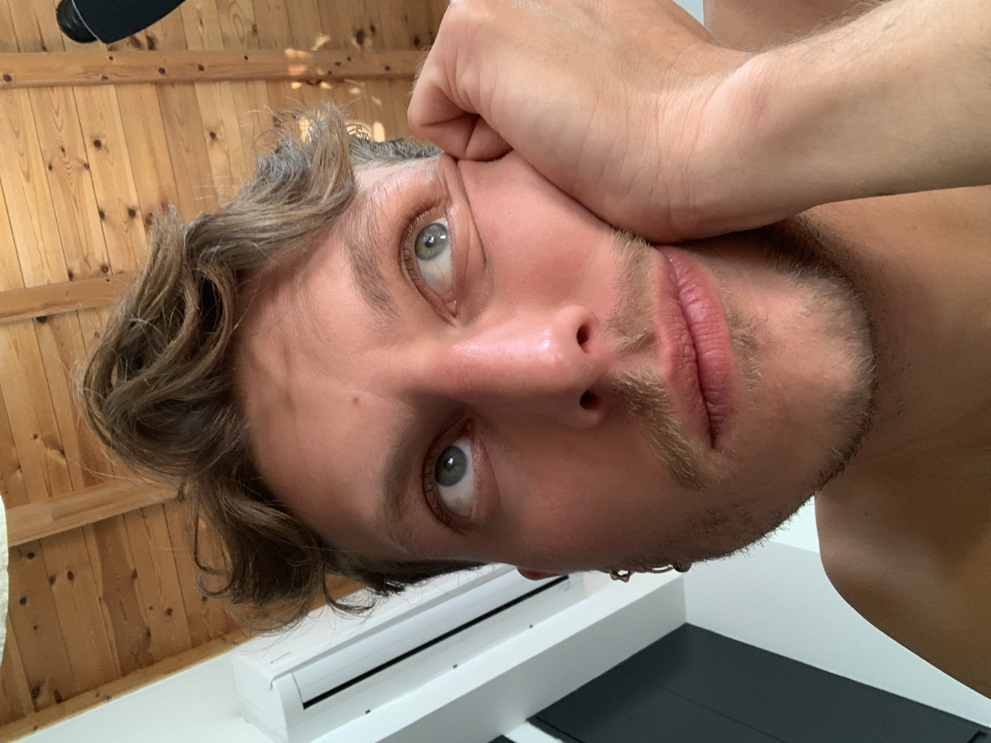 A close-up, rotated portrait shows a Caucasian male with light brown, wavy hair and stubble. His hand is near his face, appearing to support his head. He has light eyes and pink lips. In the background, there is wood paneling, an air conditioning unit and a section of a dark surface. The location, Tel-Aviv, Israel, is not discernible from the image.