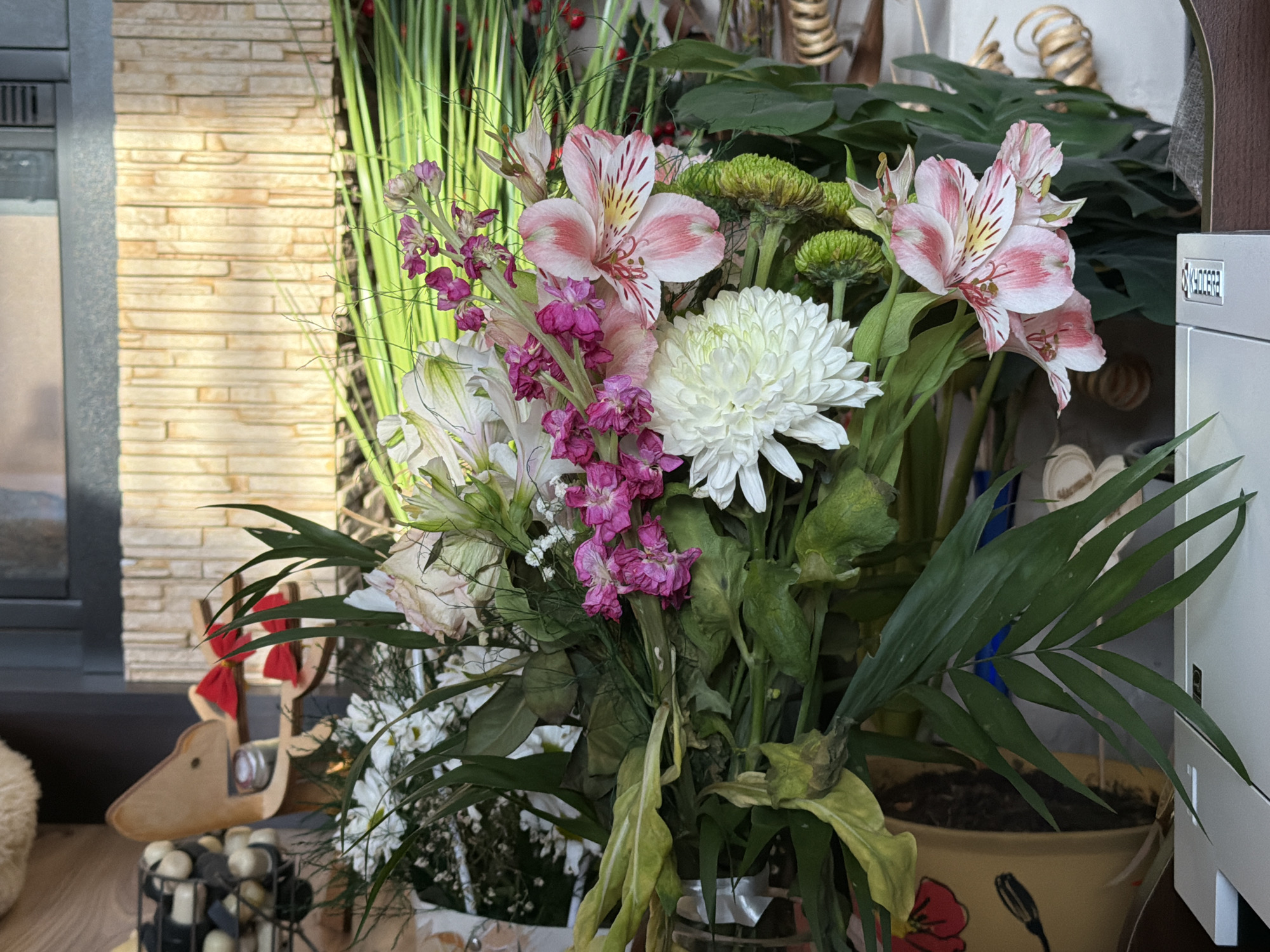 A medium-shot depicts a flower arrangement consisting of alstroemeria, chrysanthemums, and stock. The arrangement is centered in the image, with the flowers displaying shades of pink, white, and green. Behind the arrangement, there are green foliage, tall thin stems, and corkscrew willow branches. The vase is transparent, with a faint silver ribbon tied around it. To the left of the arrangement is a wooden reindeer figurine on a wooden surface, and to the right is a potted palm. The backdrop includes a stone-textured wall and a piece of office equipment. The location is Unknown, Unknown.